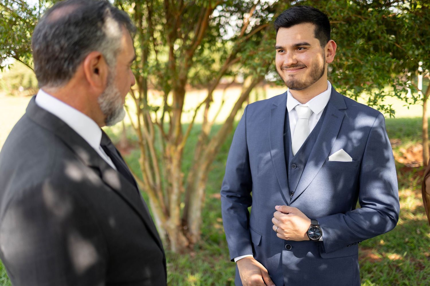 A groom in a blue suit smiles at an older man in a black suit outdoors near green trees at a wedding.