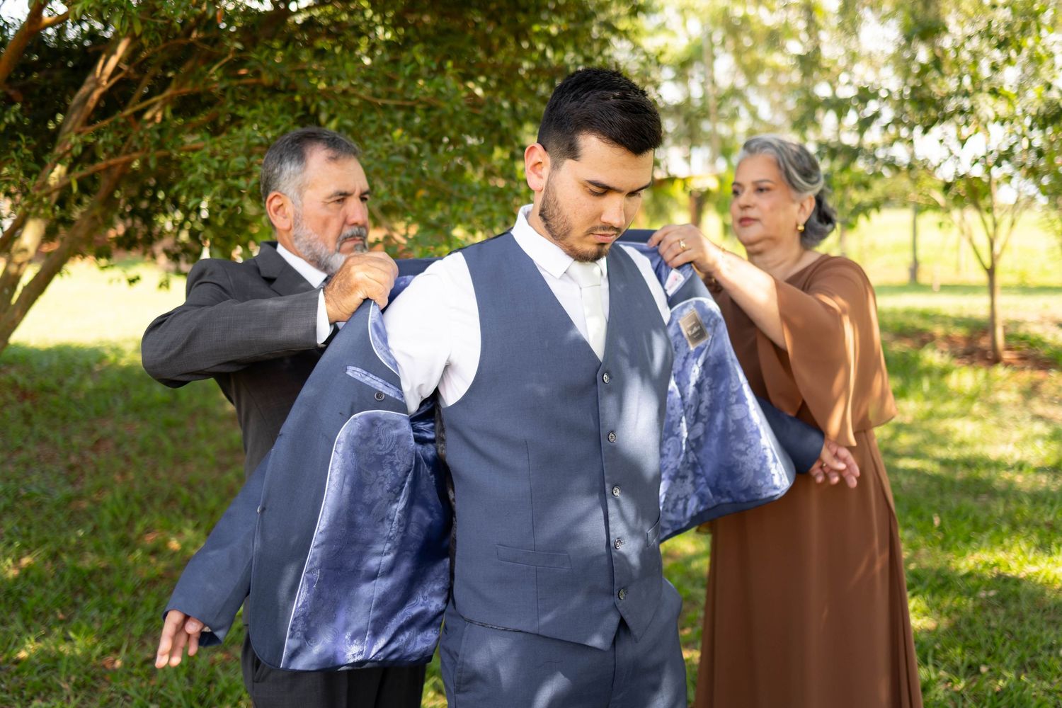 Parents helping groom put on suit jacket outdoors before wedding ceremony in green garden setting.