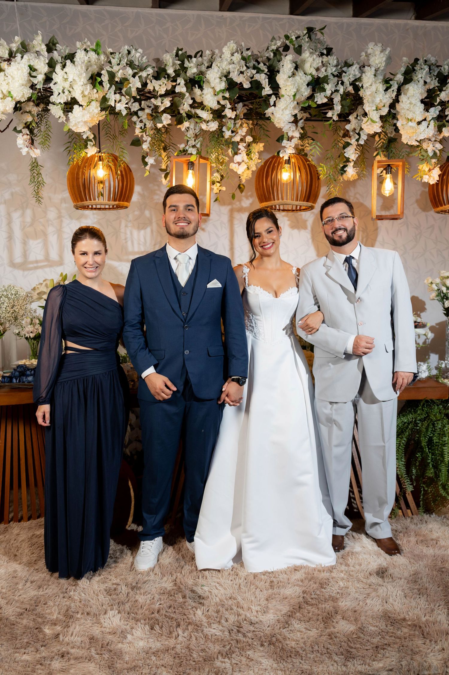 Bride and groom pose with two guests at elegant wedding reception with floral arch and hanging lanterns.