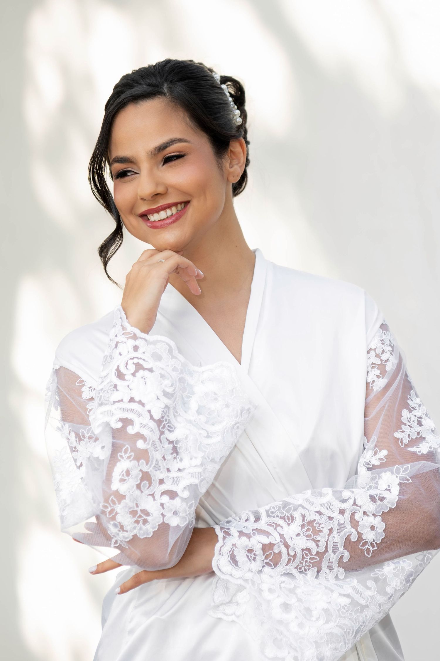 Smiling bride in white lace robe with hand on chin, posing elegantly against a light background.