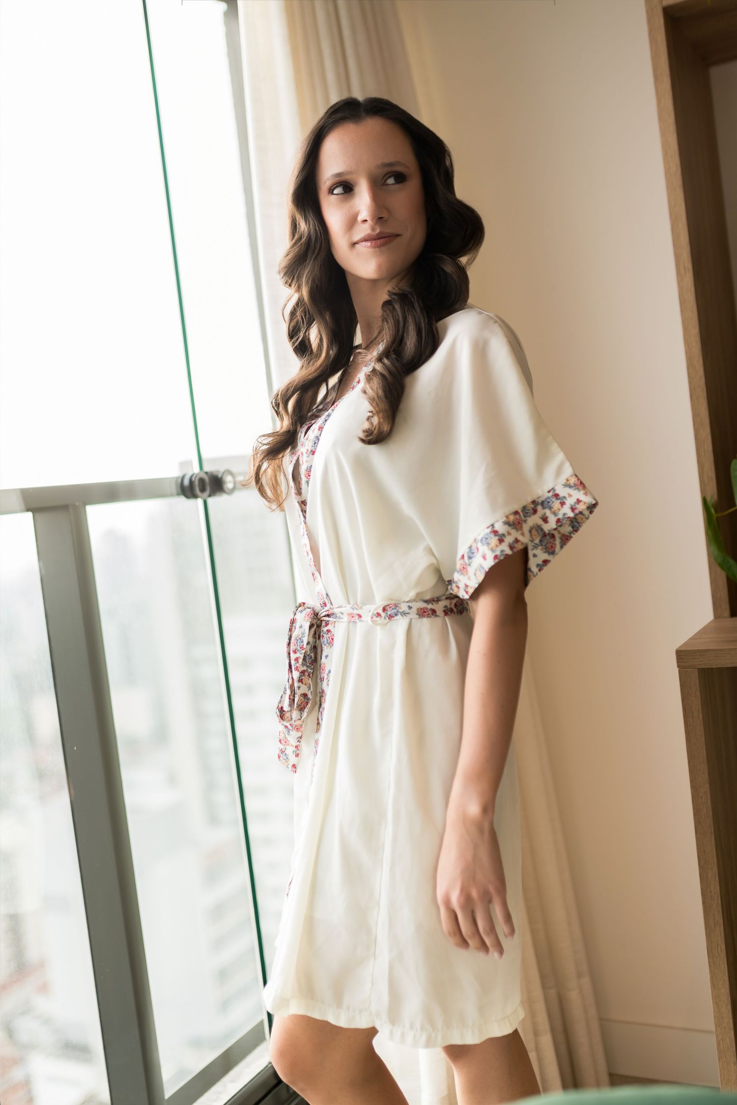 Woman in white floral-trim robe standing by large window in elegant room.