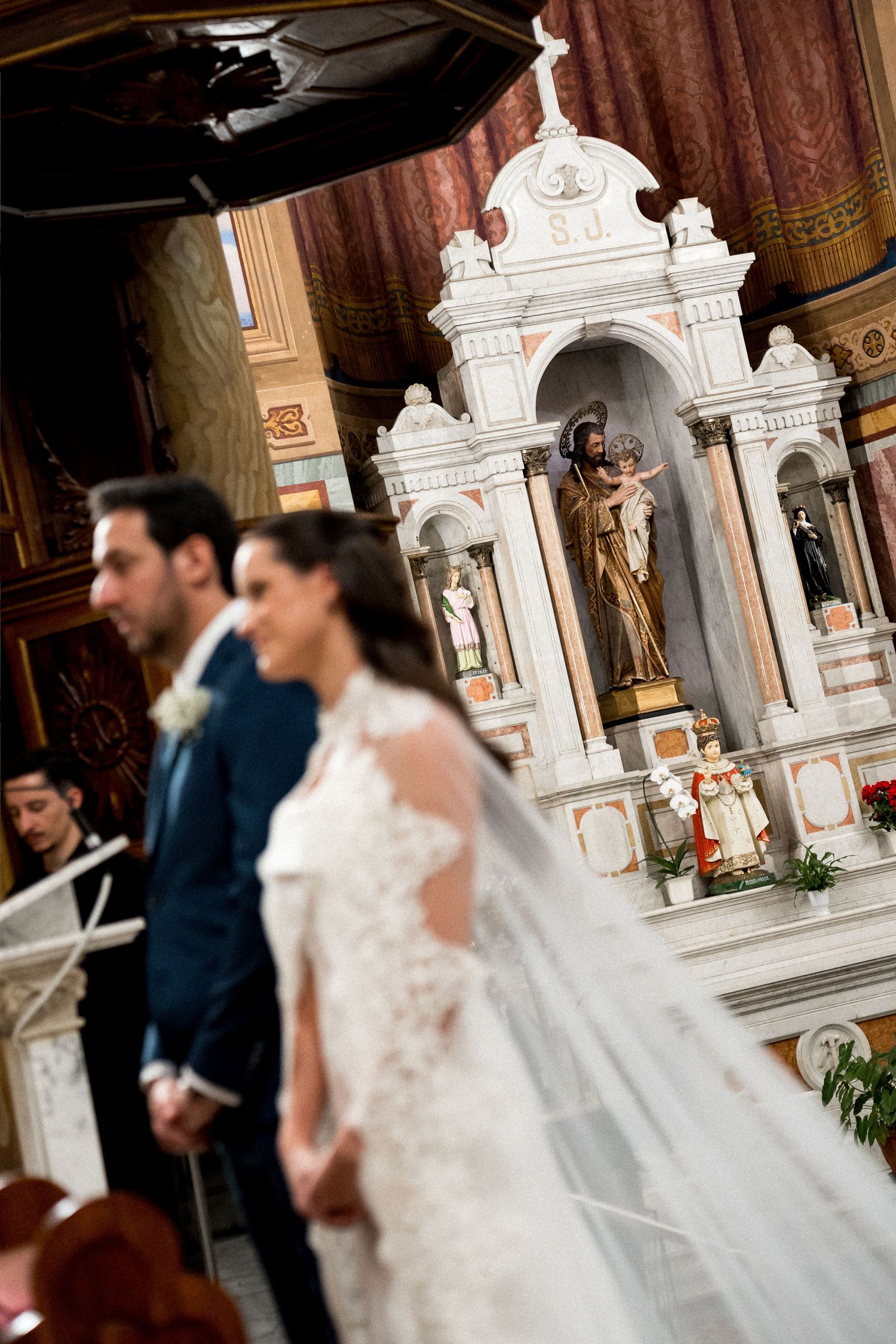 Bride and groom stand at church altar during elegant wedding ceremony with ornate white marble religious statues.