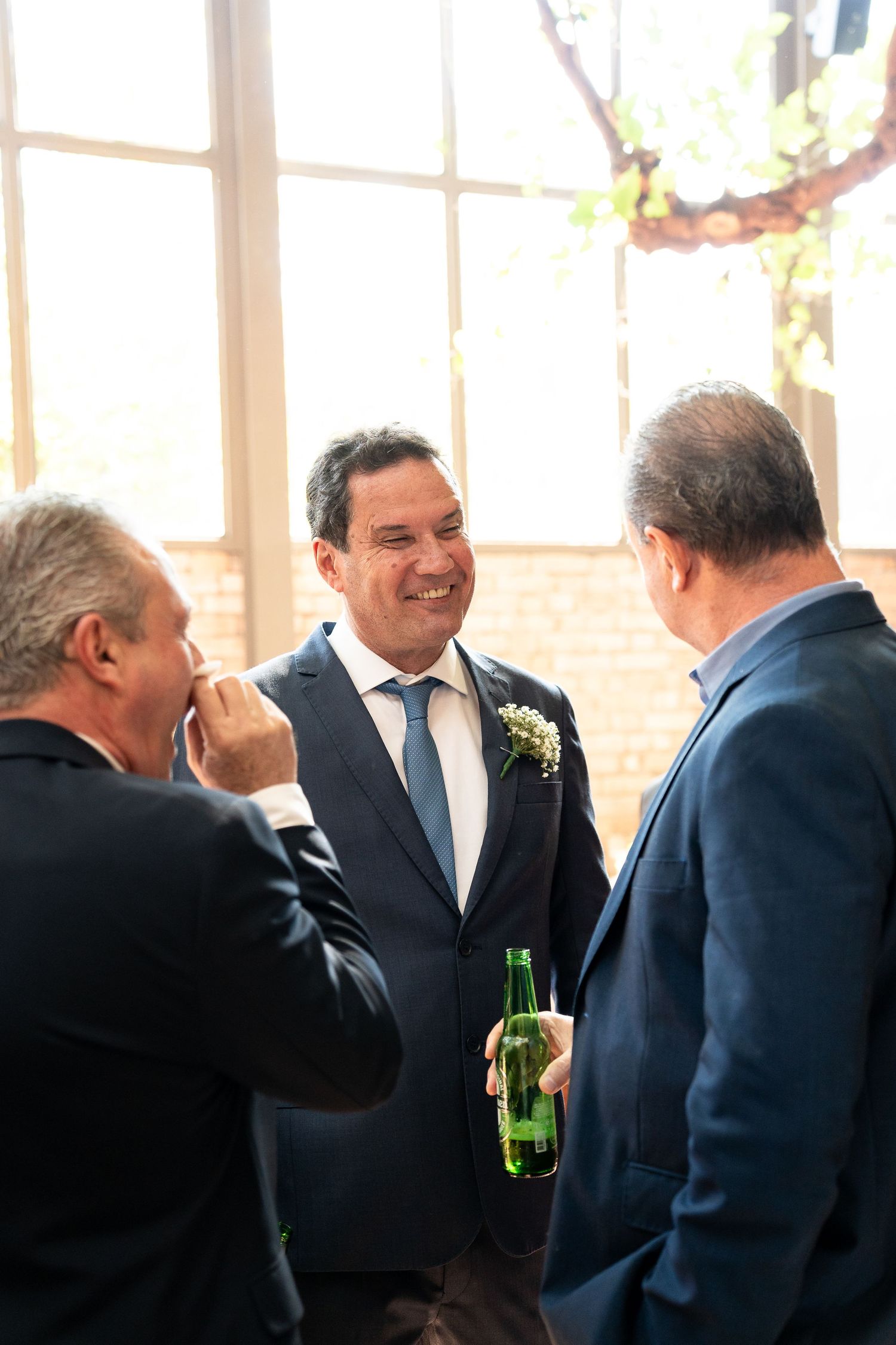 Groom in navy suit with boutonniere smiles while chatting with guests holding a green beer bottle at a wedding reception.