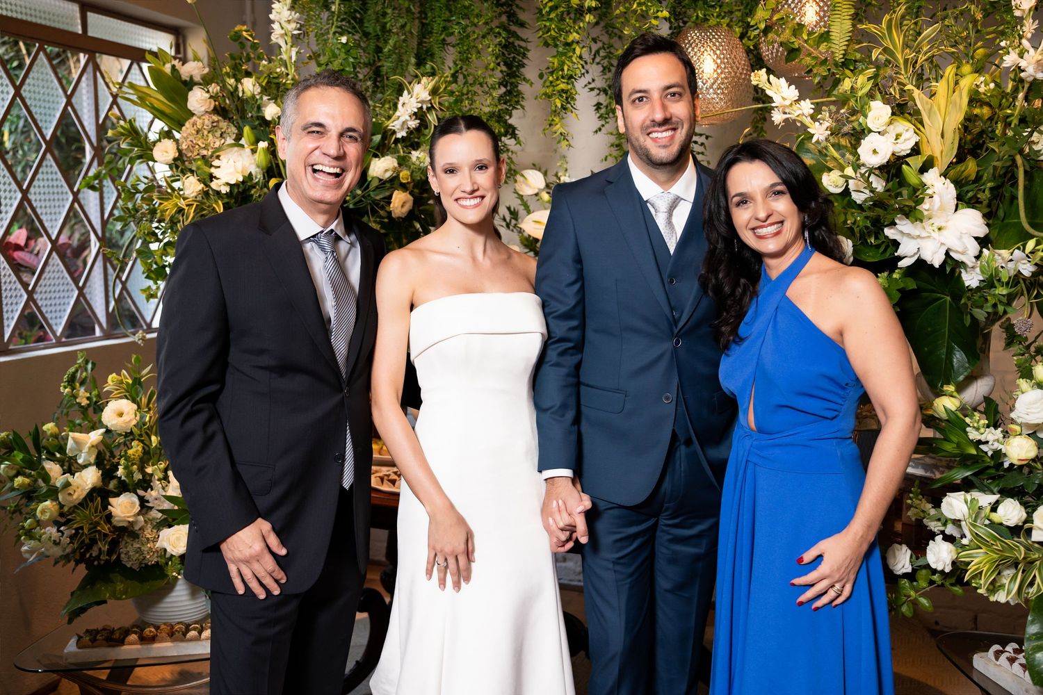 Two couples pose together at a wedding, bride in white gown, groom in navy suit, guests in blue dress and gray suit.