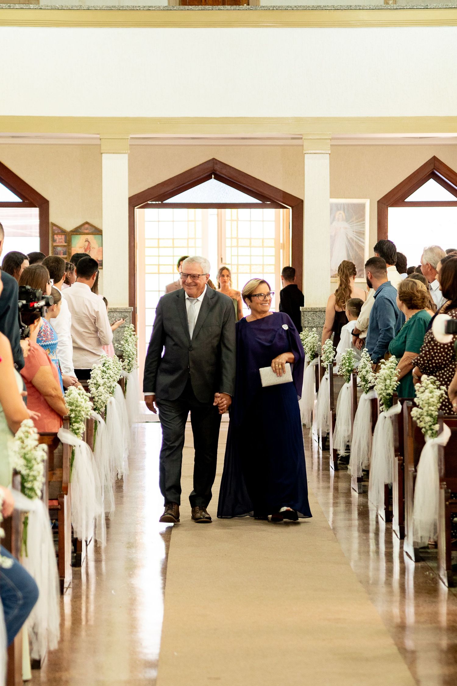 Elderly couple walking down church aisle at wedding ceremony, surrounded by guests and floral pew decorations.