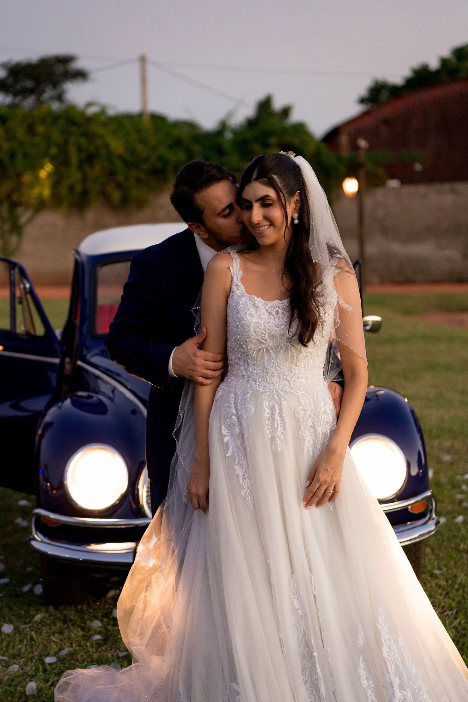 Bride in white lace gown and groom in navy suit share a romantic kiss beside a vintage blue car at dusk.