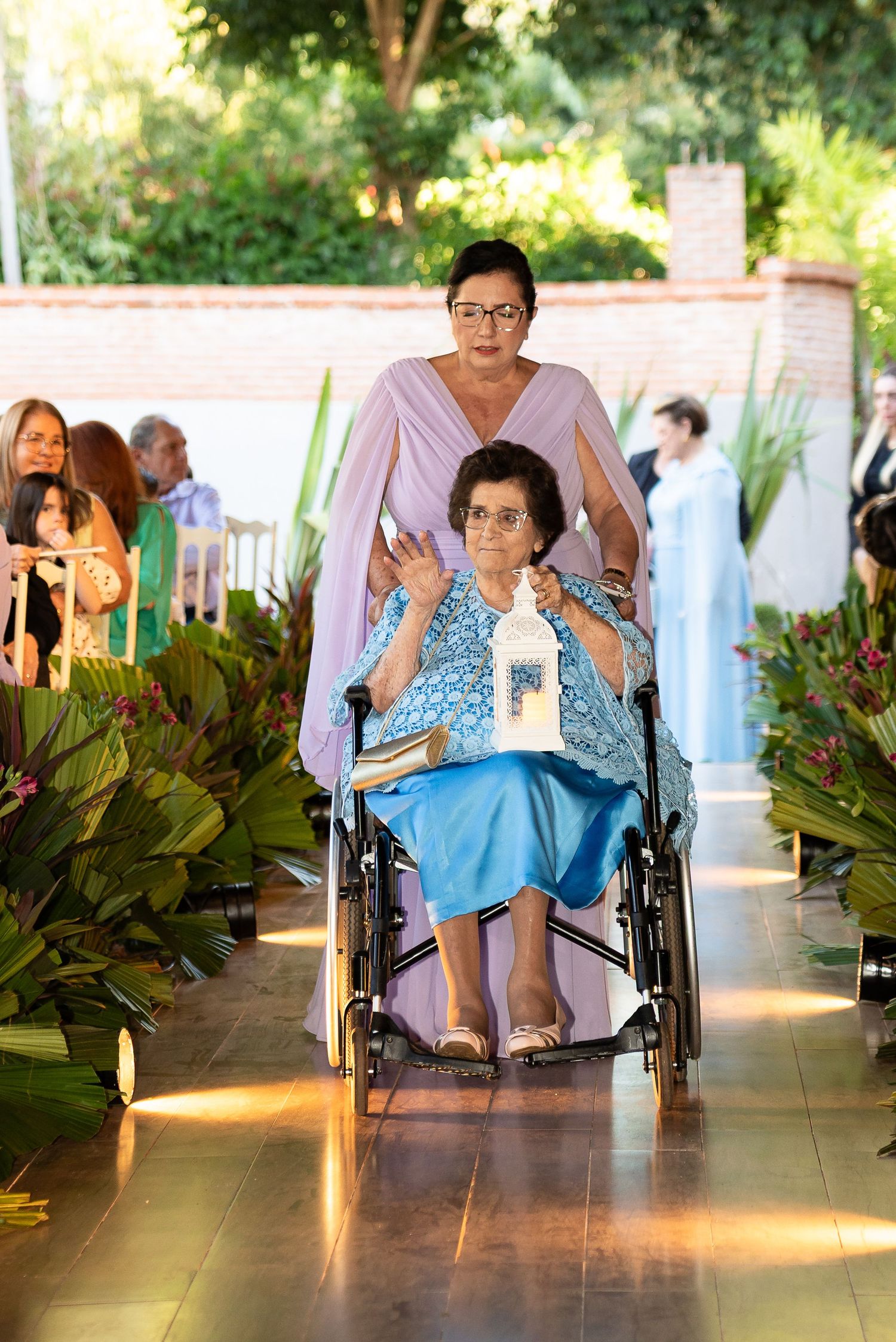 Elderly woman in blue dress being pushed in wheelchair down floral aisle at outdoor wedding ceremony.