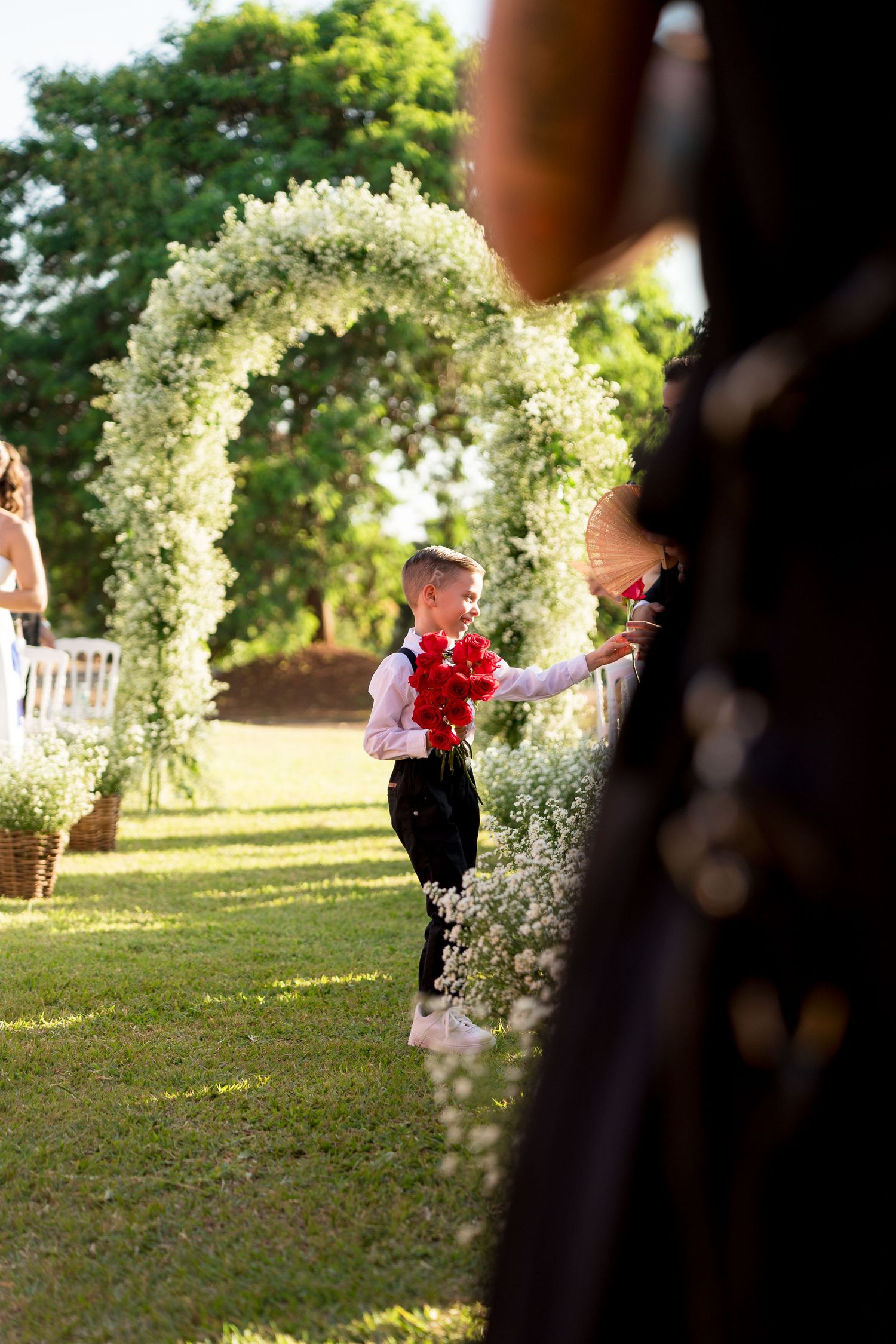 Young ring bearer in black pants holds red flowers at outdoor wedding ceremony with white floral arch on green lawn.