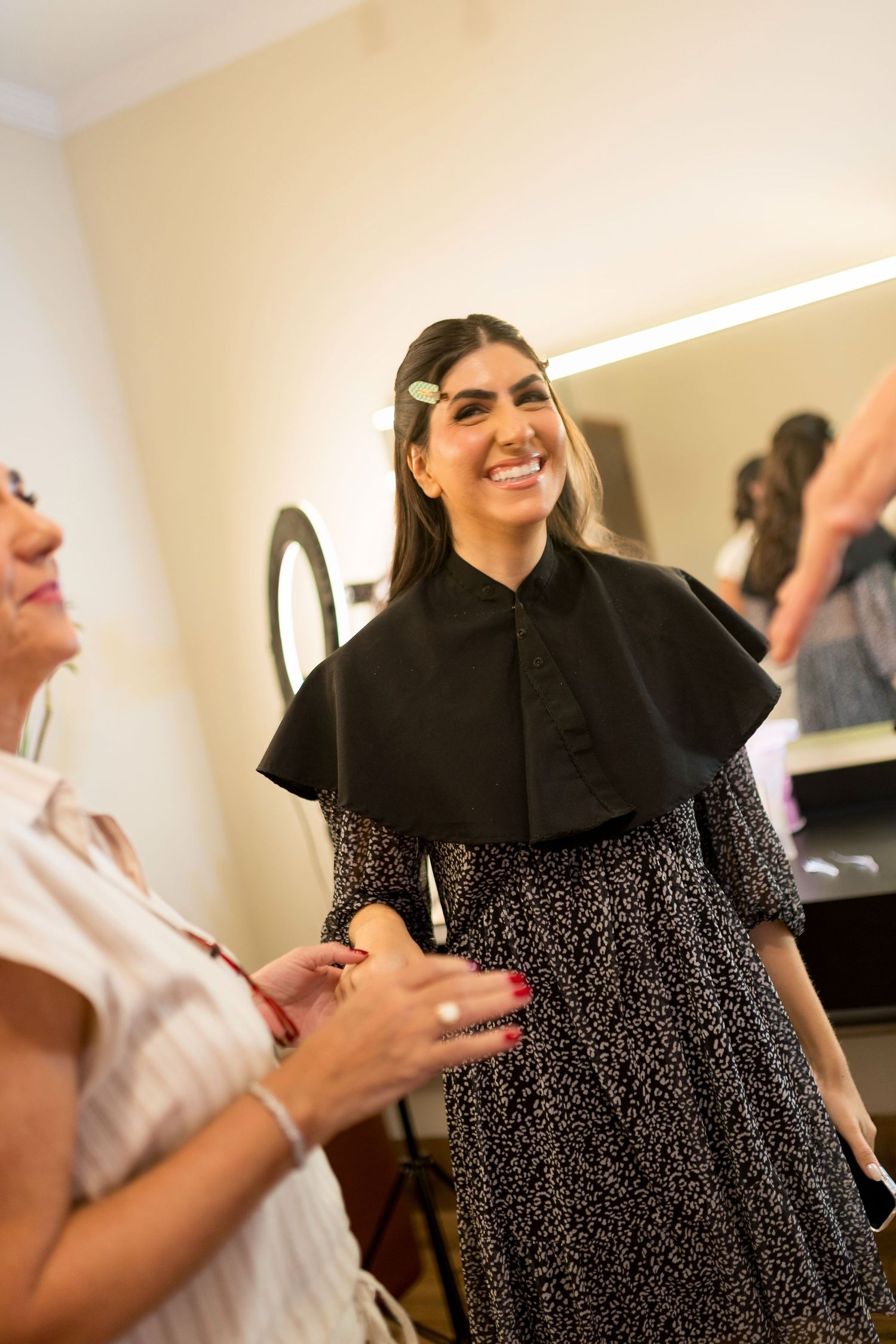 A smiling woman in a salon wearing a black cape and hair clips, chatting with another woman during a hair appointment.