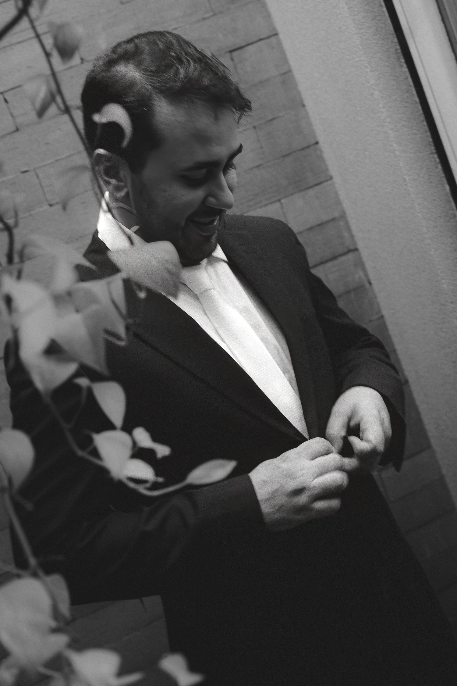 Black and white photo of a smiling groom in a tuxedo adjusting his jacket near a leafy plant.