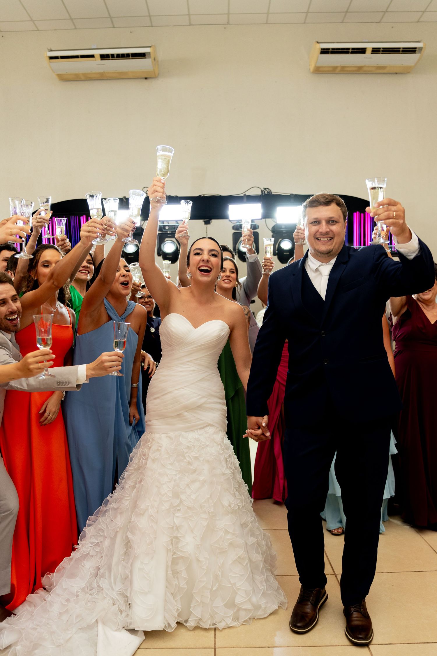 Bride and groom toast champagne glasses at wedding reception, surrounded by cheering guests in colorful formal attire.
