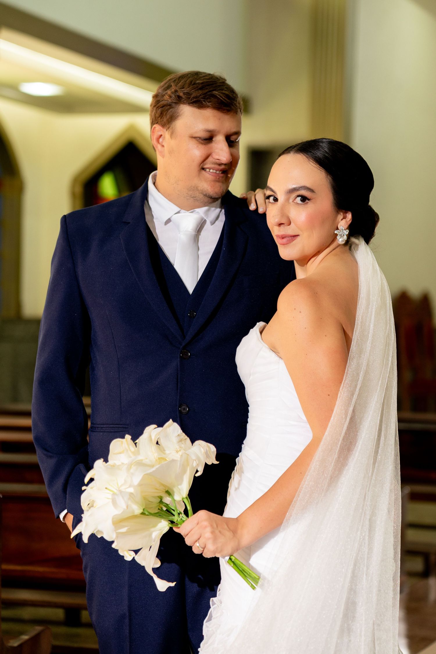 Bride and groom posing in church, bride holding white calla lily bouquet wearing veil, groom in navy suit.
