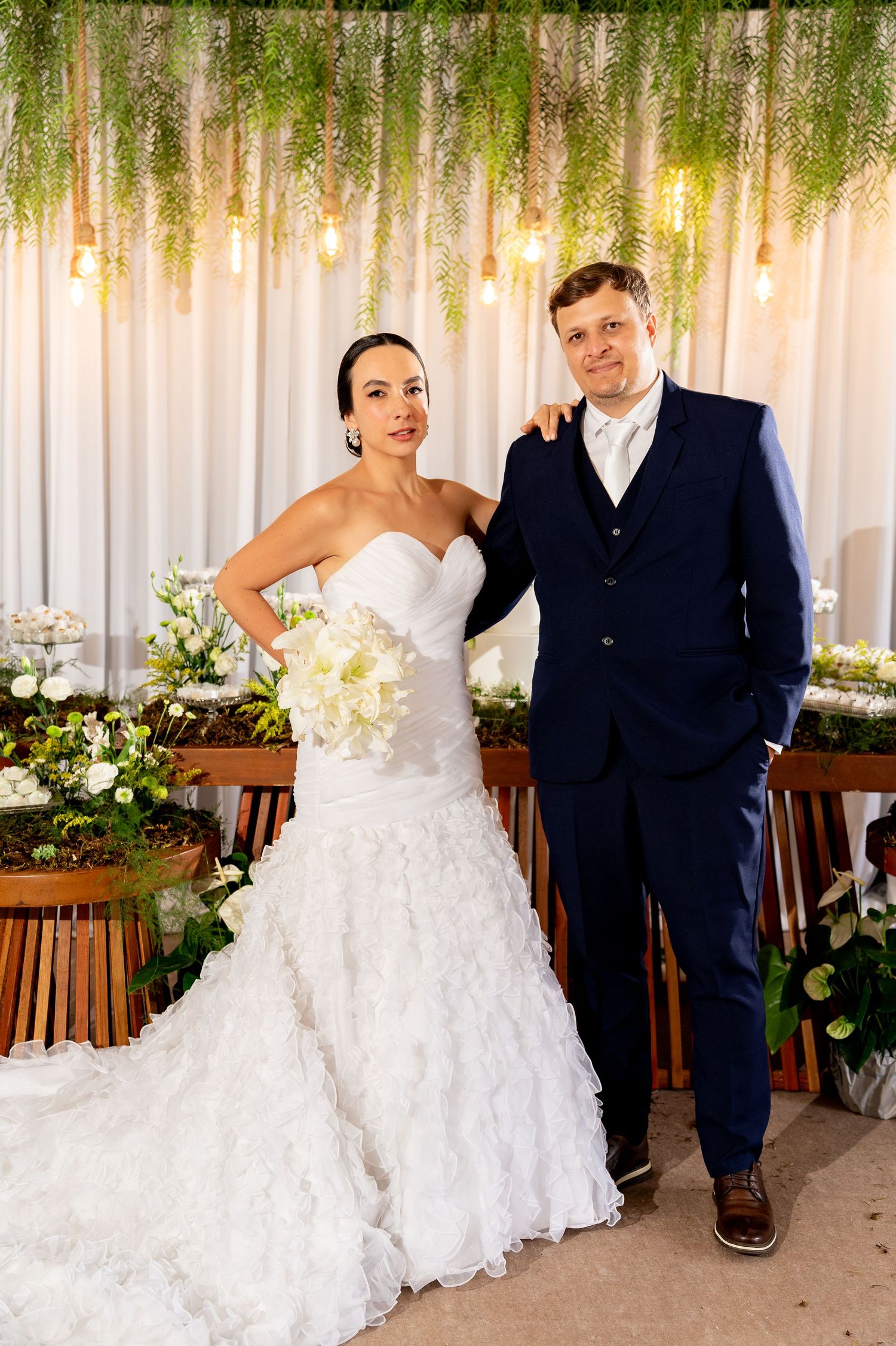 Bride in white ruffled gown and groom in navy suit pose together at elegant floral wedding ceremony backdrop.