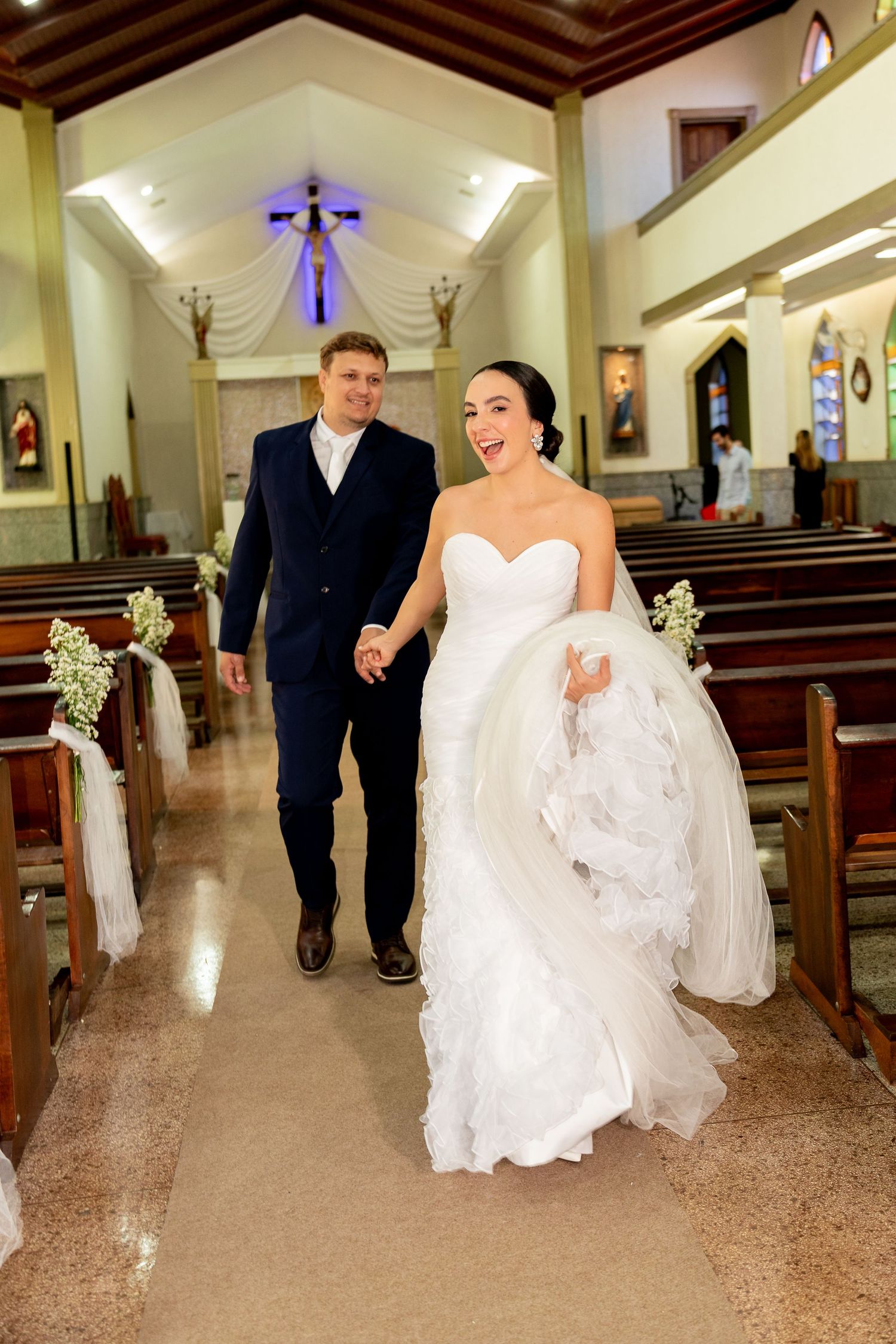 Joyful bride and groom walking down church aisle after wedding ceremony, bride in white strapless gown holding dress.