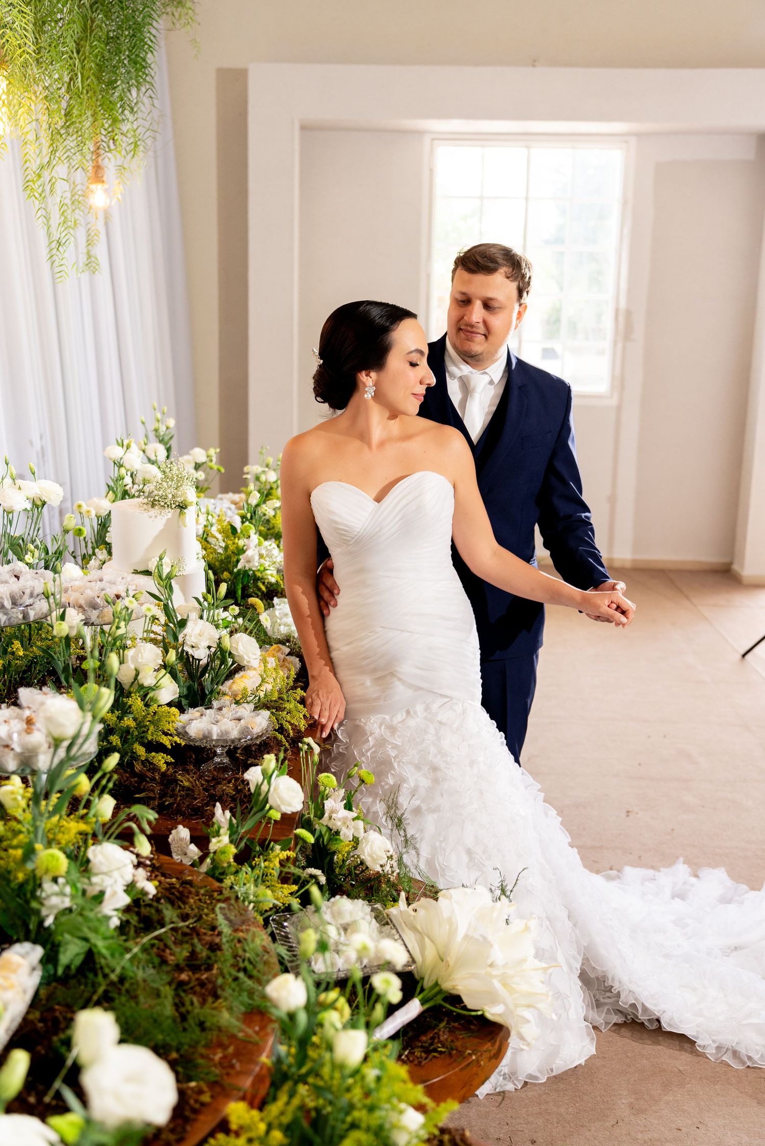 Bride in strapless mermaid gown and groom in navy suit hold hands beside lush white floral arrangements indoors.