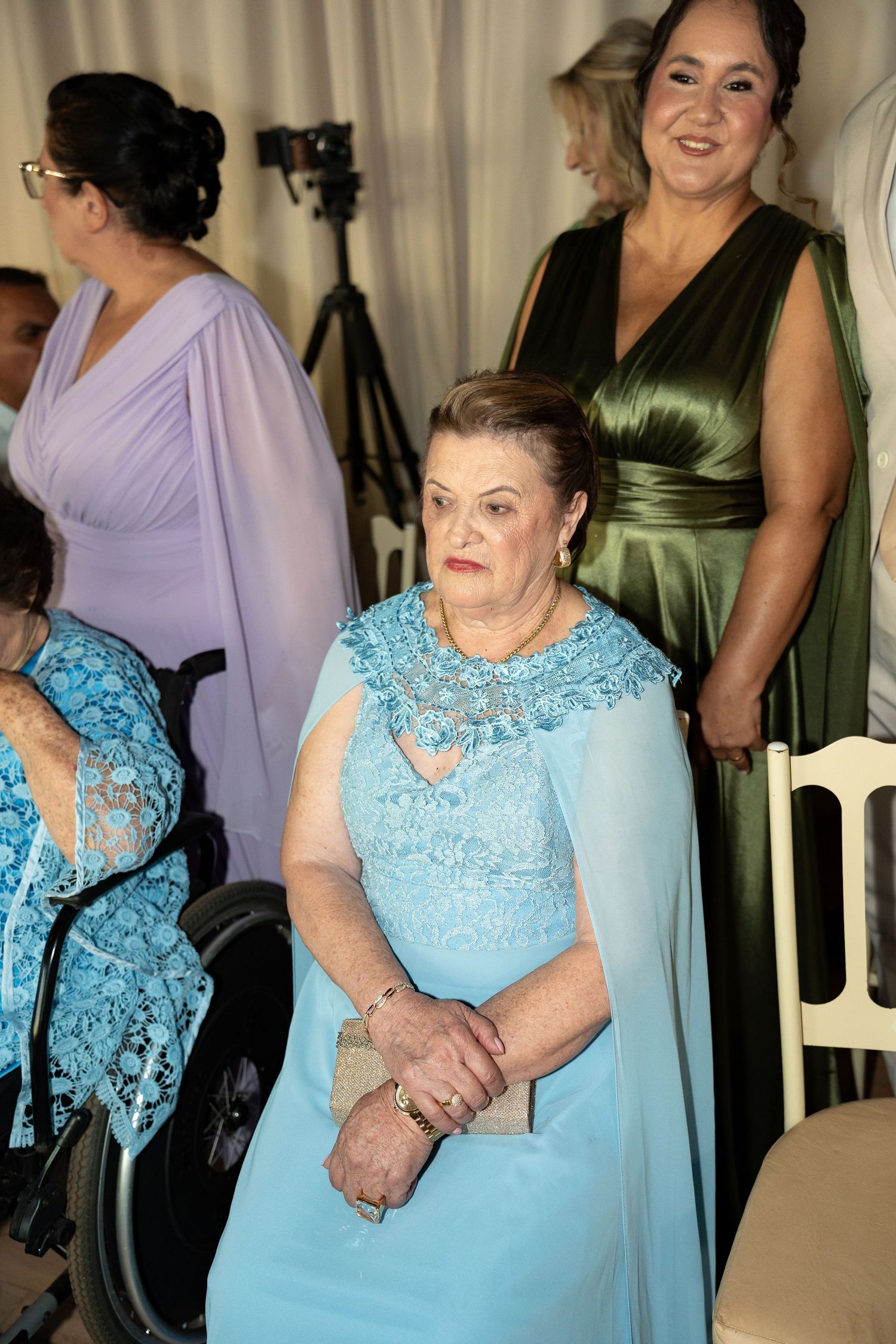 Elderly woman in light blue lace gown sits at formal event, with guests in lavender and olive green dresses behind her.