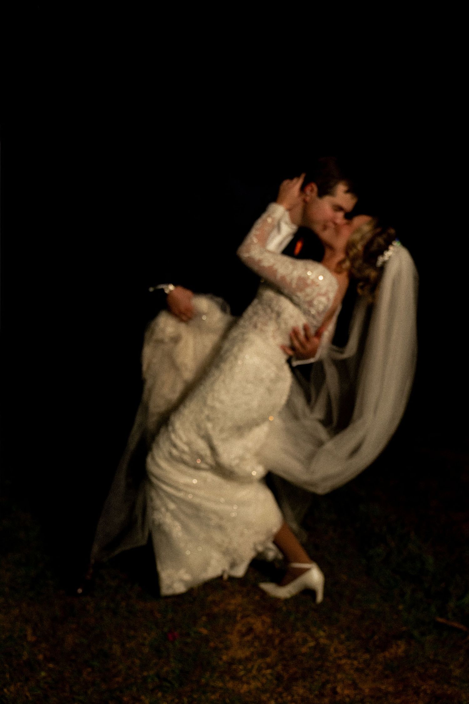 Bride and groom sharing a romantic dip kiss outdoors at night, bride in lace gown with veil flowing.