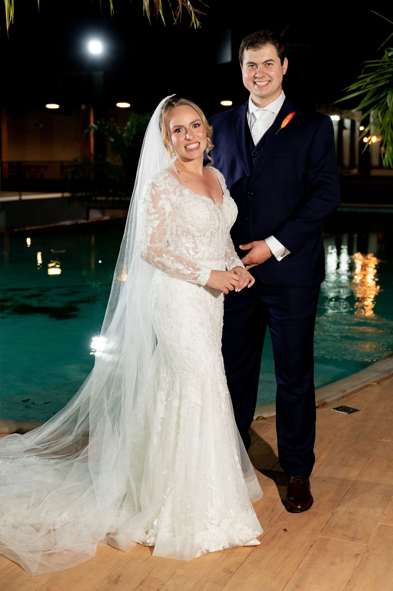Bride in lace gown with veil and groom in navy suit pose by illuminated pool at night wedding venue.