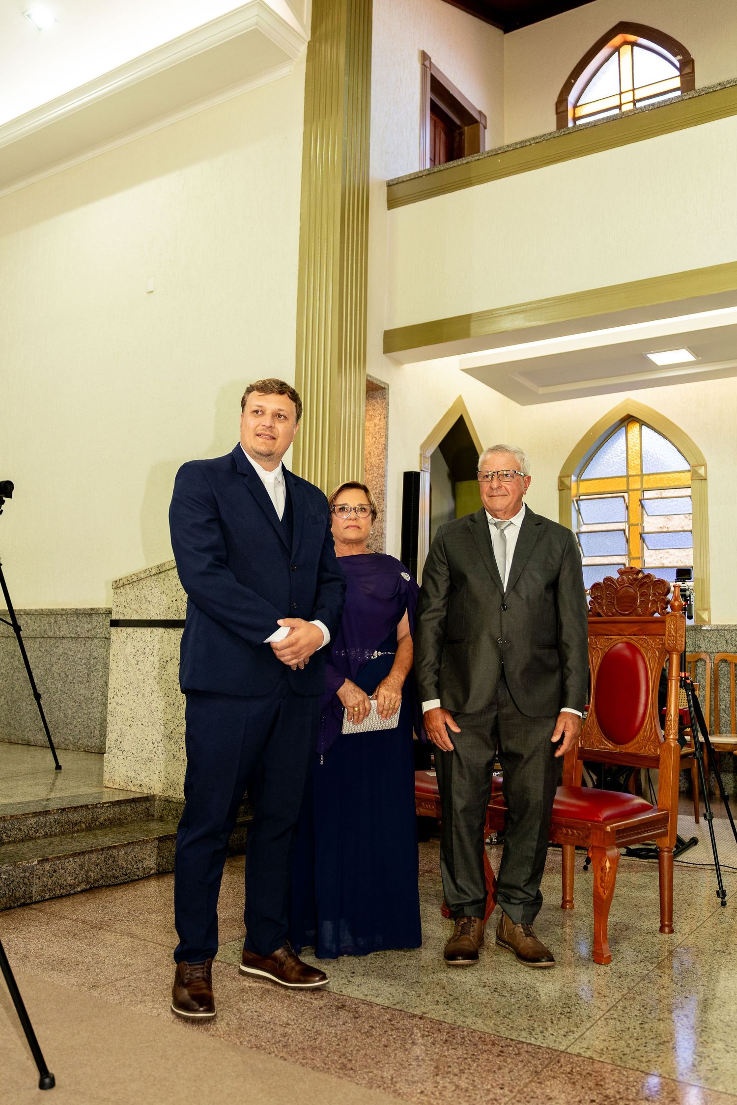 Three formally dressed people posing together inside a church with a cross and ornate wooden chair visible behind them.