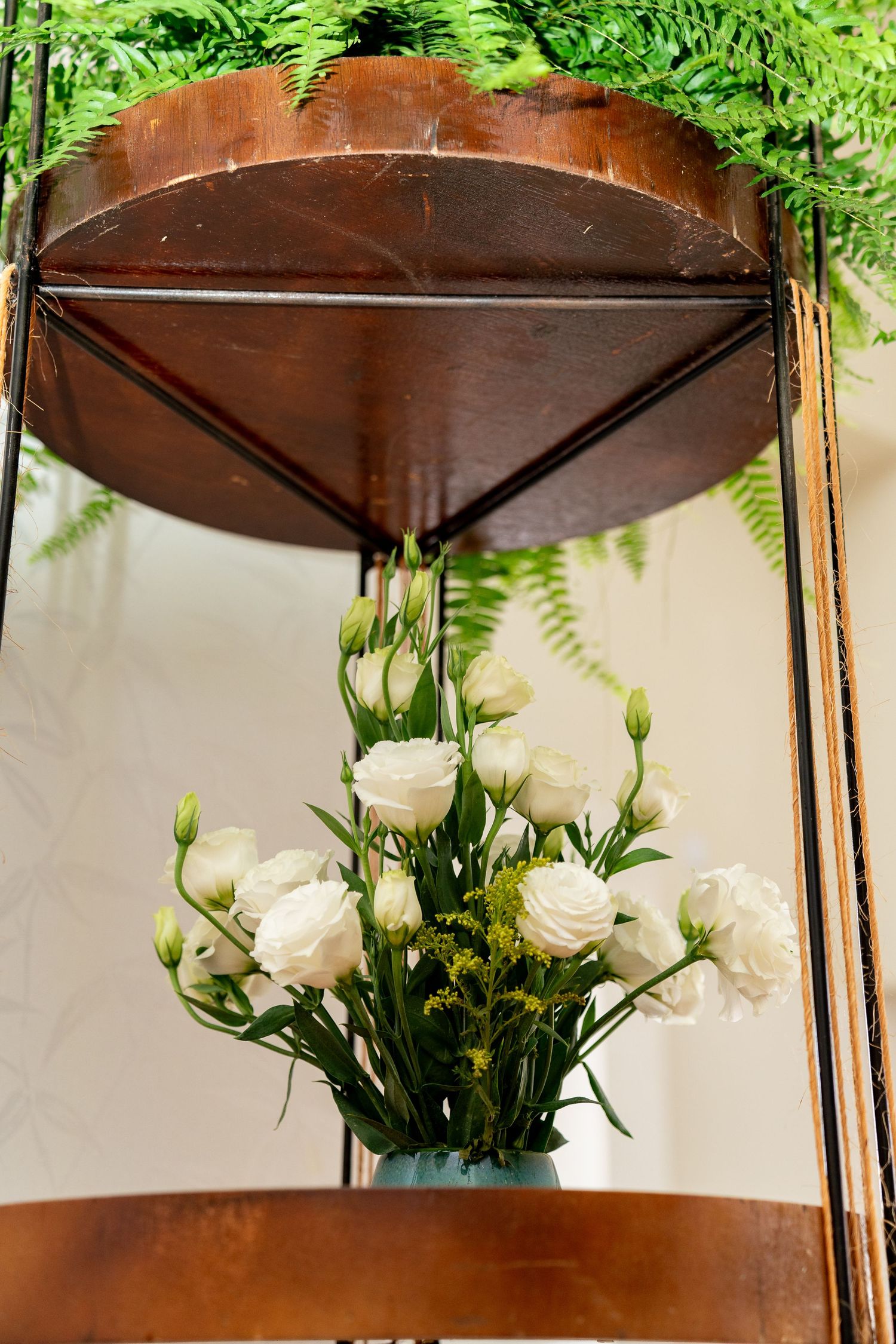 White roses bouquet in teal vase displayed under rustic wooden shelf with fern greenery backdrop.
