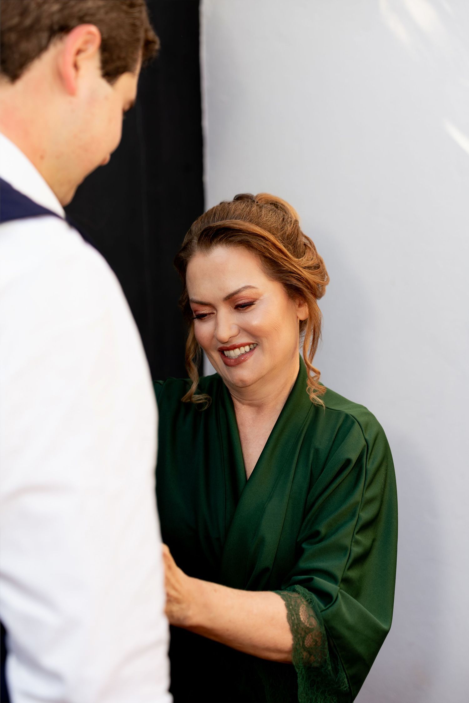 Smiling woman in green satin robe with lace trim laughing with a man in a white shirt during wedding preparations.