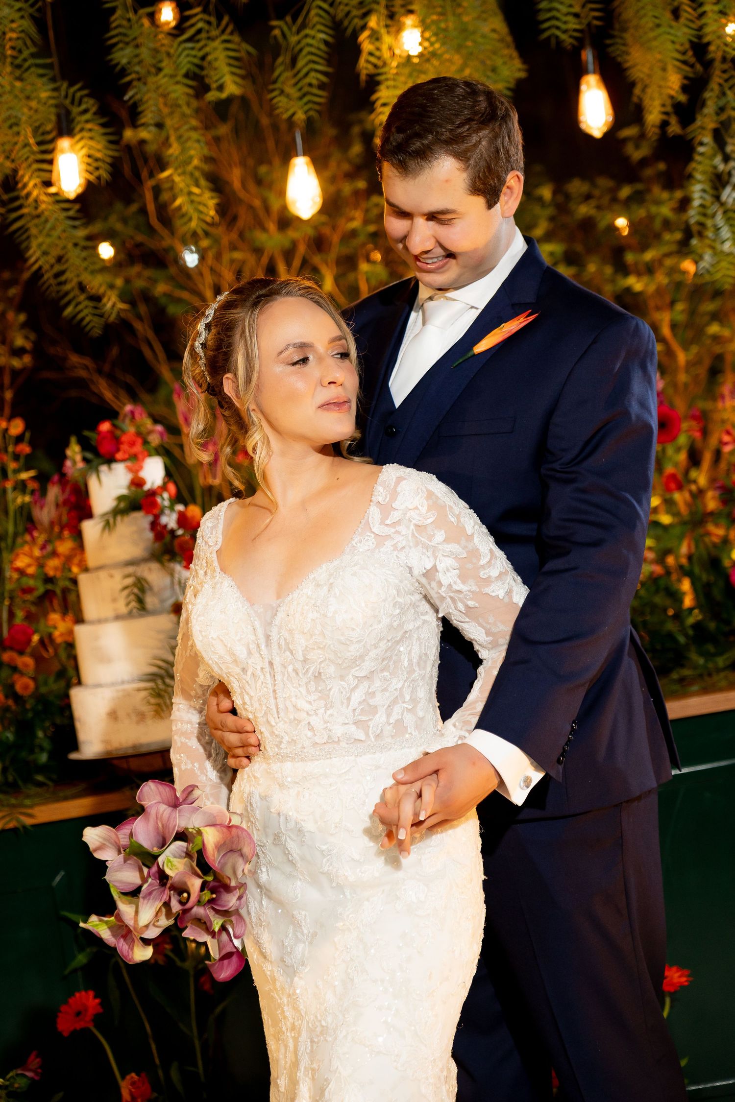 Bride in lace gown holds flowers as groom in navy suit embraces her before floral backdrop with string lights.
