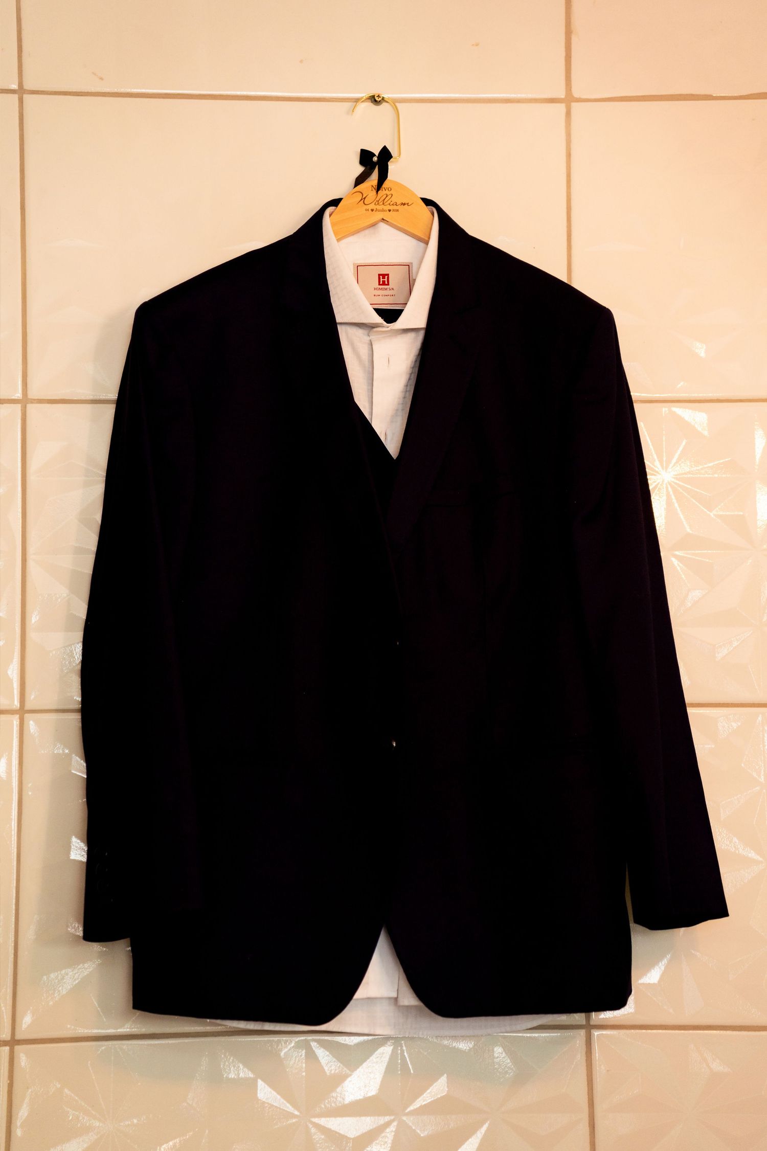 Black suit jacket on wooden hanger displayed against white tile wall with white dress shirt visible.