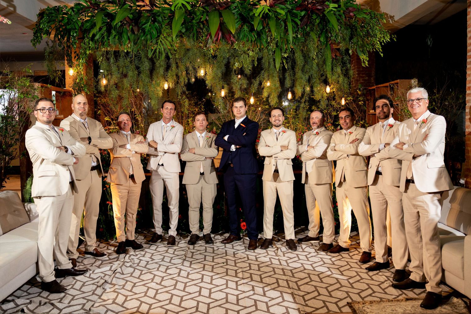 Groom in navy suit poses with groomsmen in tan suits under lush greenery at elegant wedding venue.