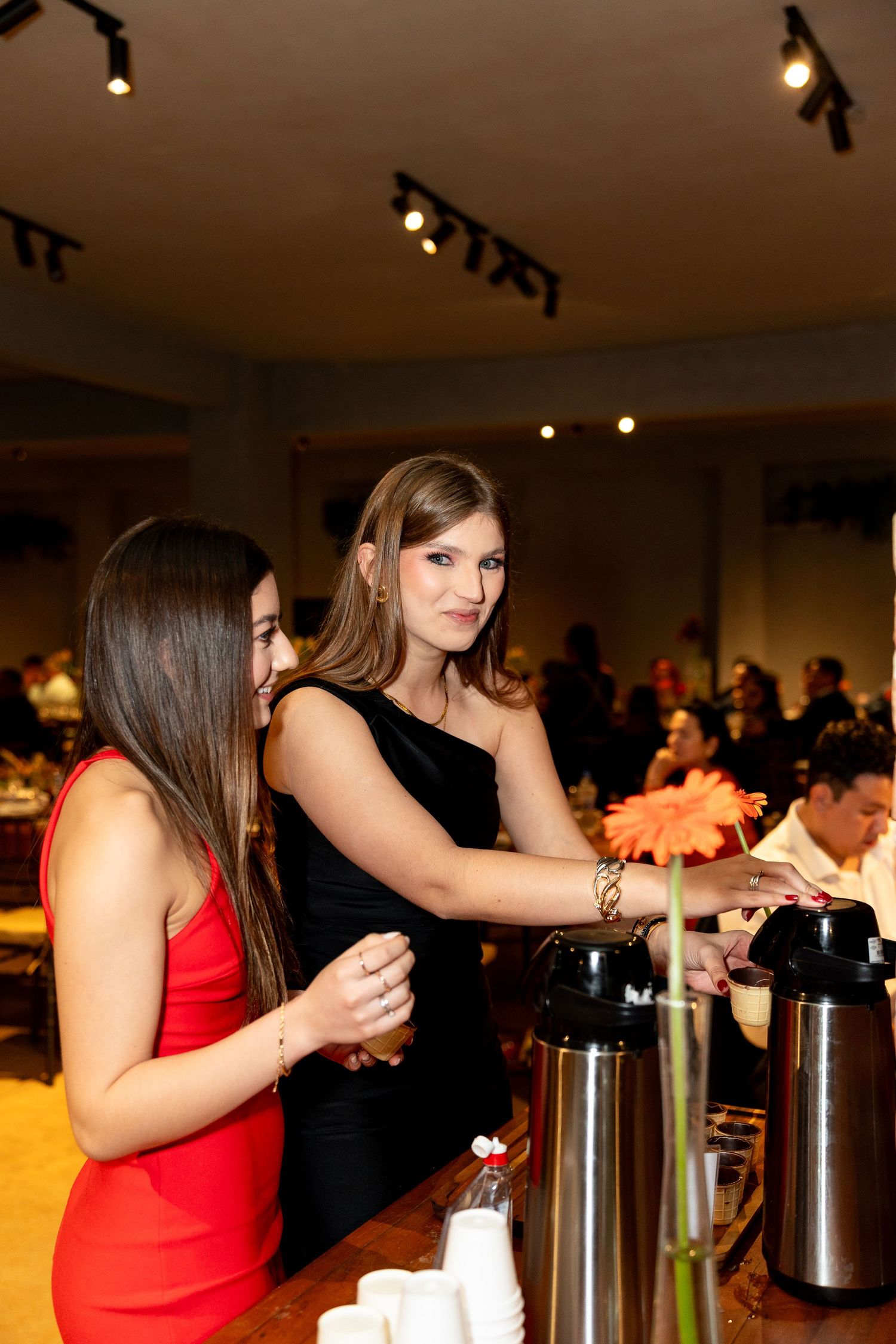 Two elegant women in red and black dresses serving drinks at a formal event bar.