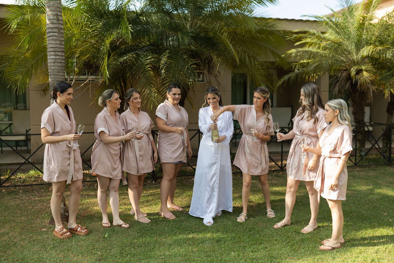 Bride in white robe with bridesmaids in tan robes celebrating outdoors near tropical palm trees.