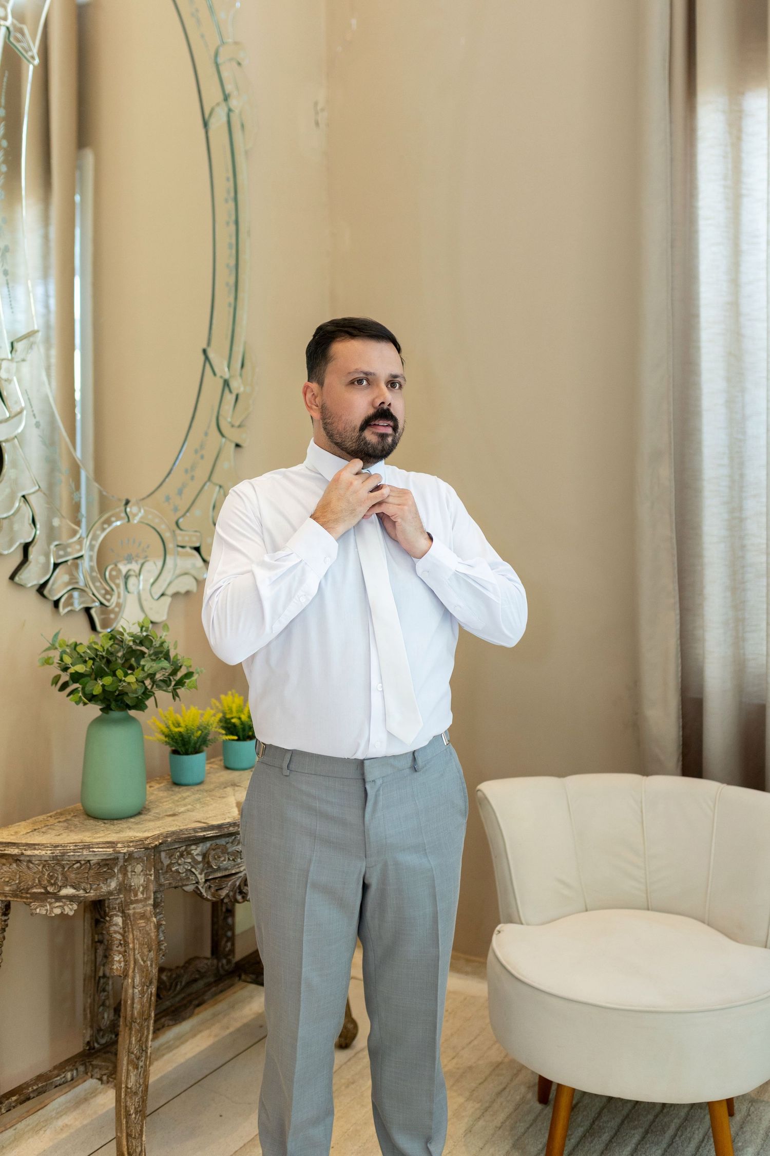 Man in white dress shirt and gray pants adjusting collar near elegant mirror and console table in stylish room.