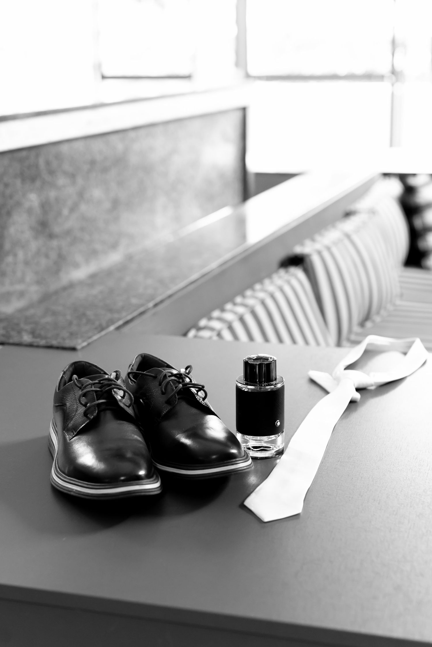 Black and white photo of mens dress shoes, cologne bottle, and white tie arranged on a table.