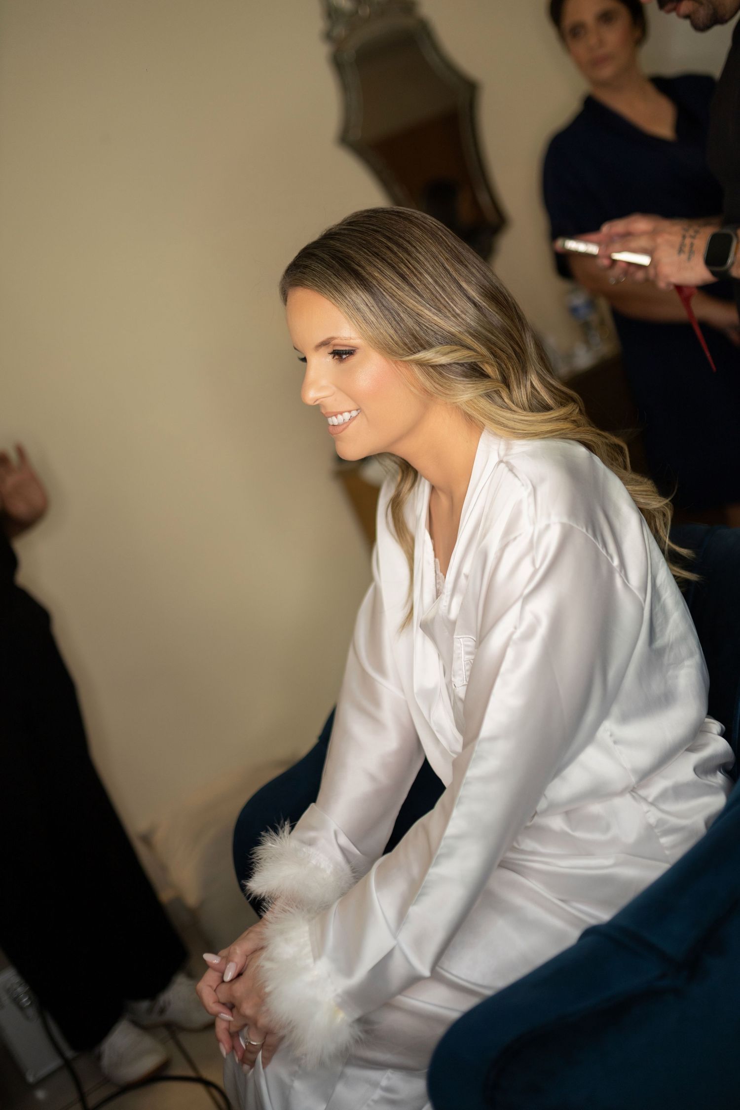 Smiling bride in white satin robe with feather cuffs sits while hairstylist works behind her during wedding preparations.