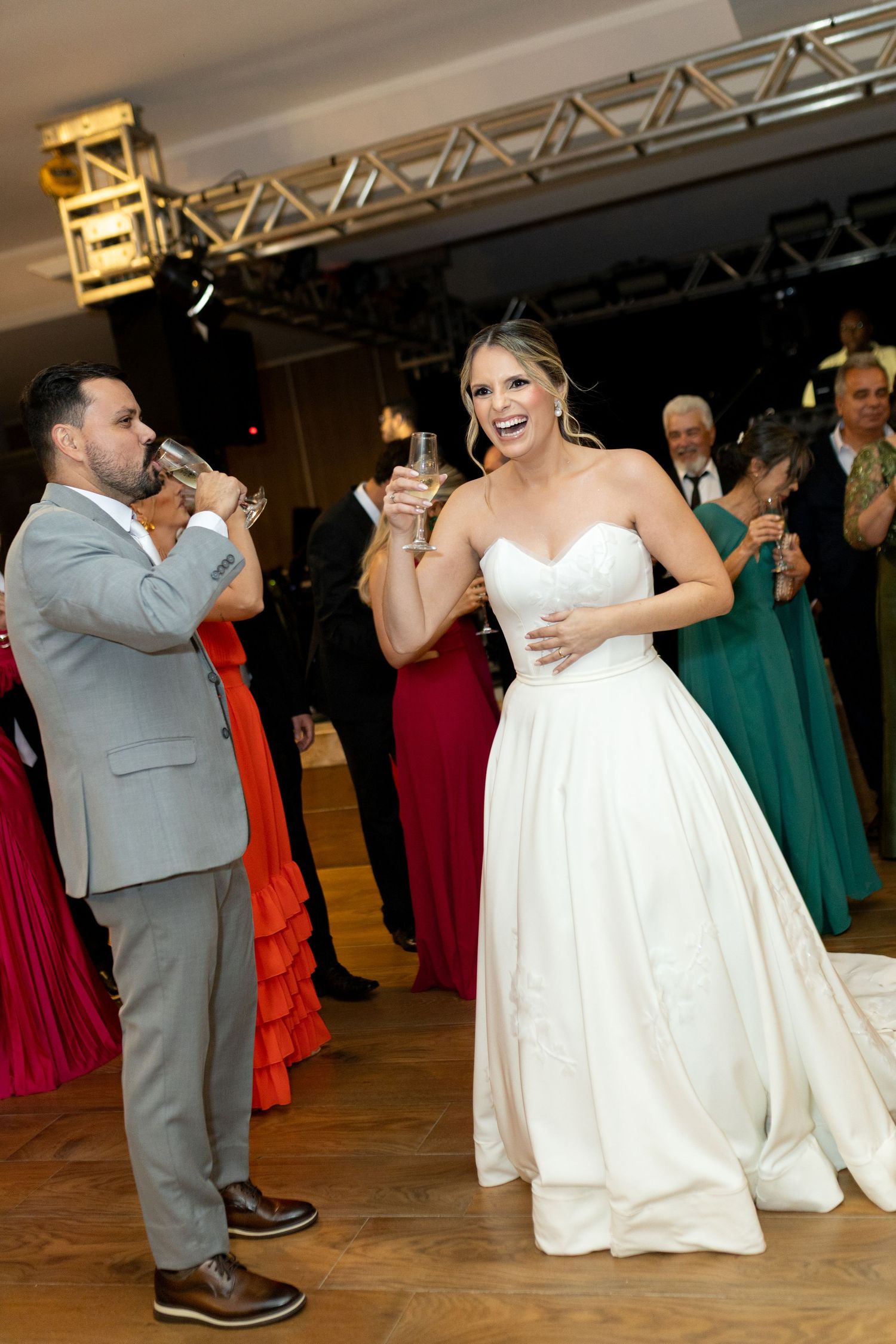 Bride in white gown and groom in gray suit toasting champagne glasses at wedding reception on dance floor.