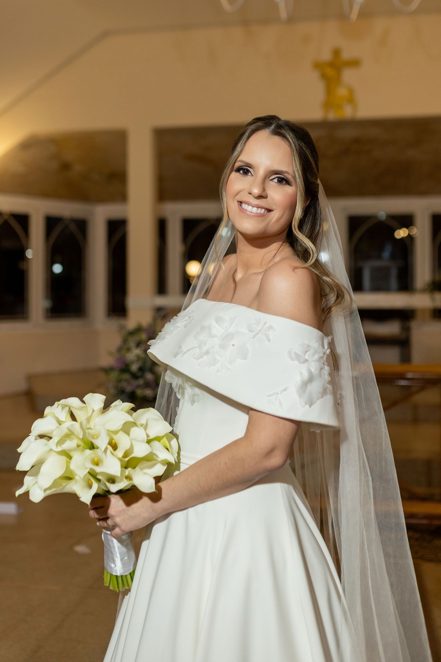 Smiling bride in off-shoulder white gown holding calla lily bouquet with veil in elegant church setting.
