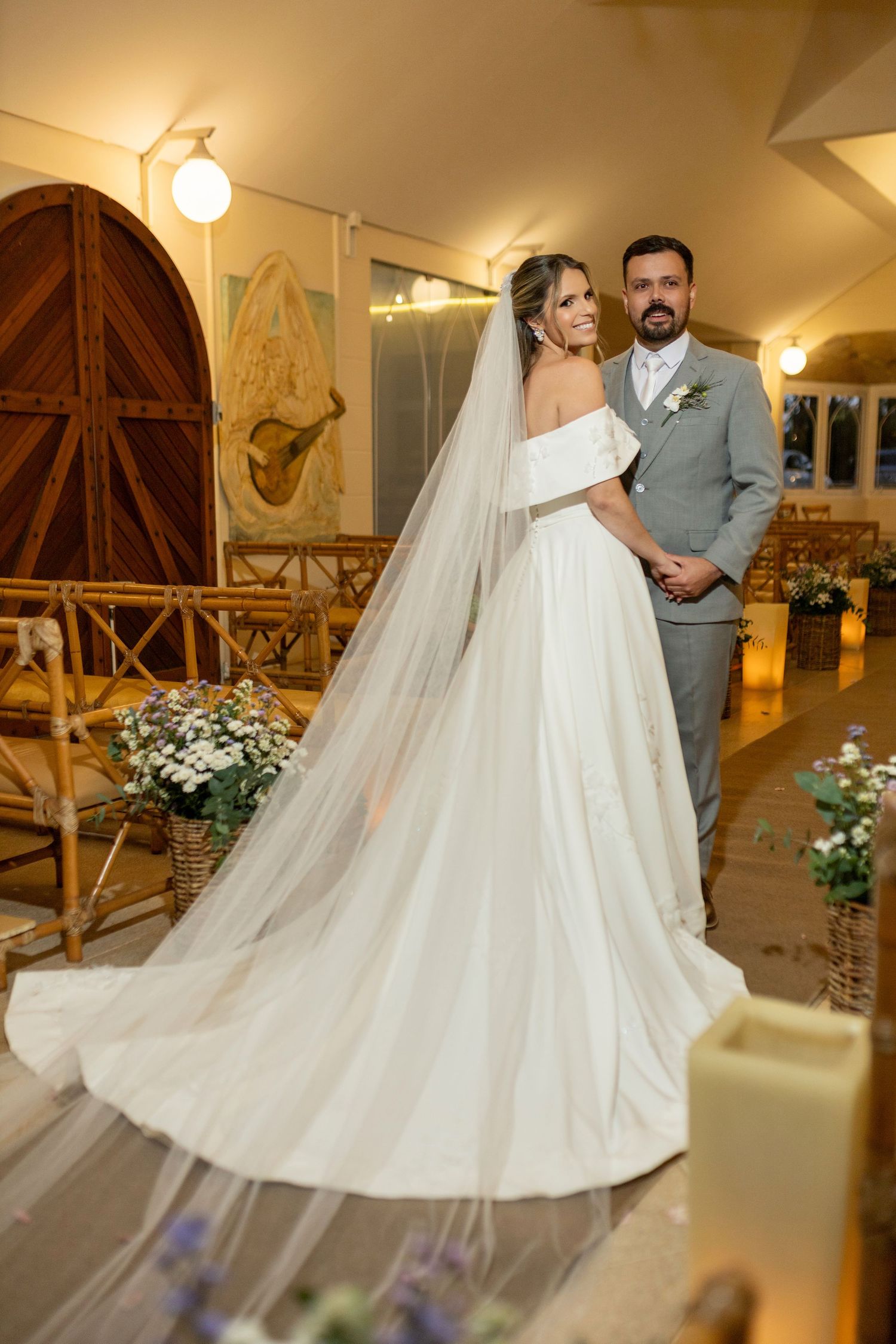 Bride in off-shoulder gown with long veil stands beside groom in gray suit inside a warmly lit chapel.