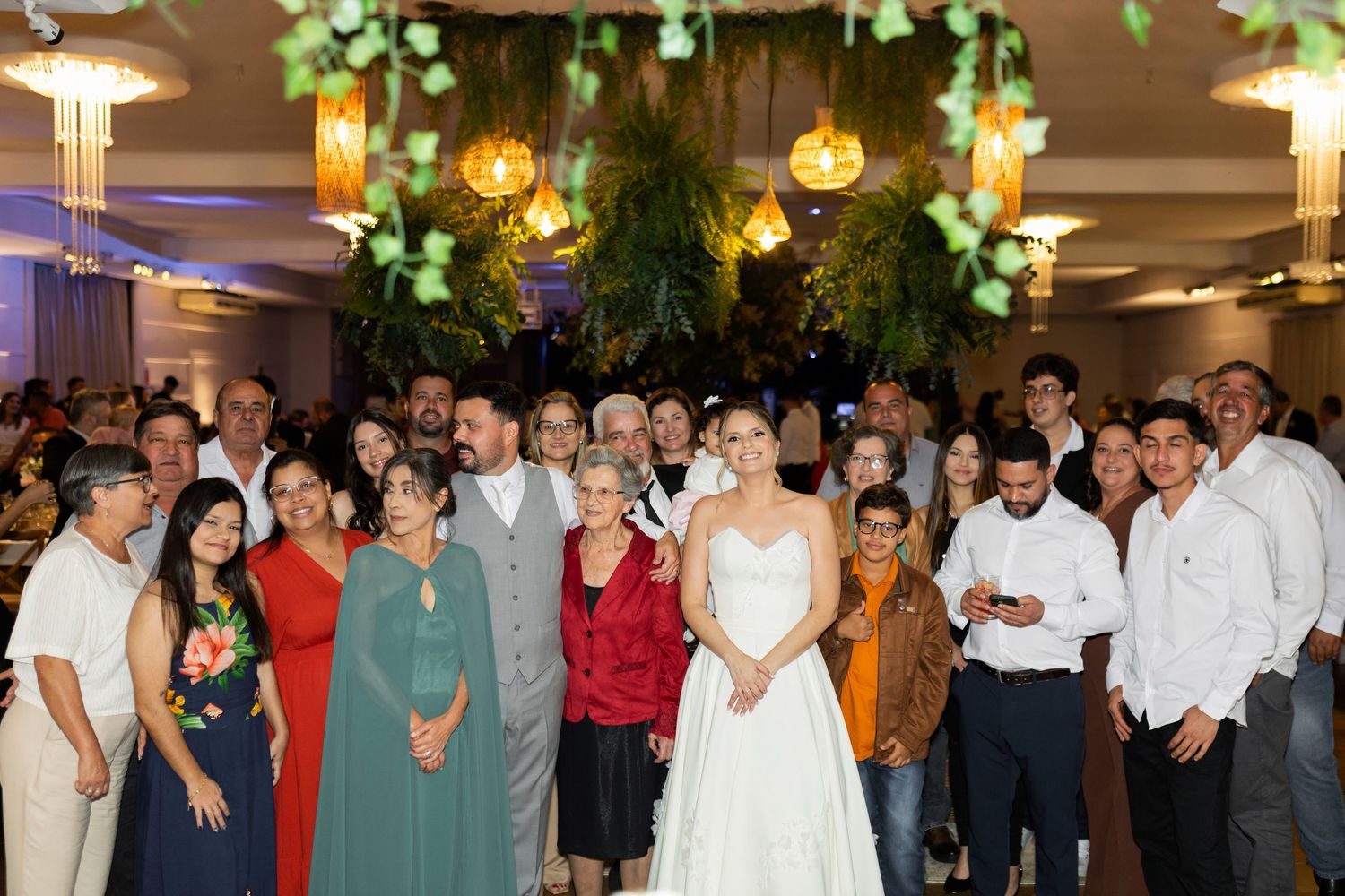 Wedding reception group photo with bride in white gown surrounded by family and friends under lush green floral ceiling decor.