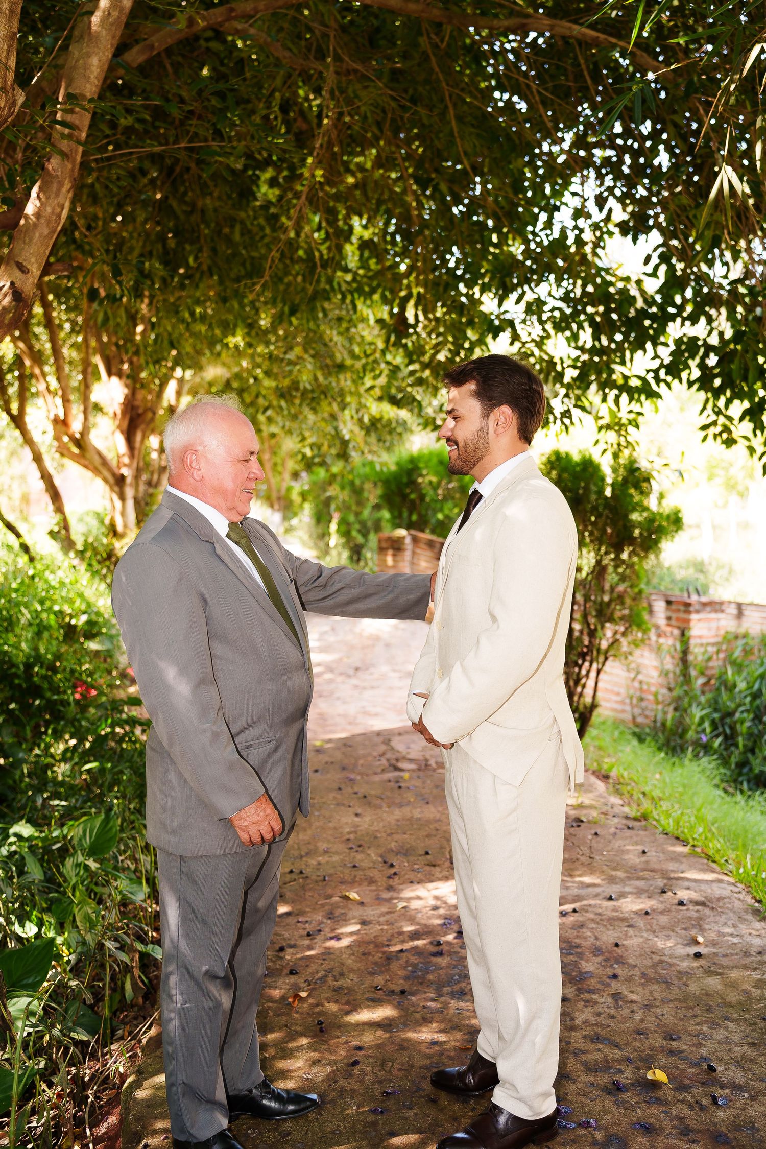 Two men in suits share a heartfelt moment outdoors, one in gray and one in ivory, likely at a wedding ceremony.