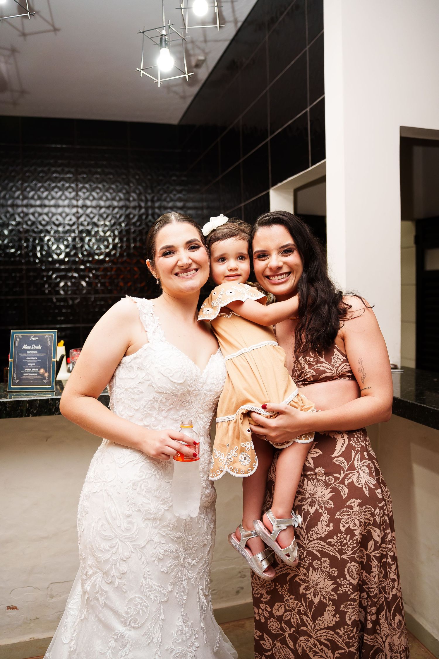 Bride in white lace gown poses with woman in floral dress holding a young girl in yellow dress at wedding reception.