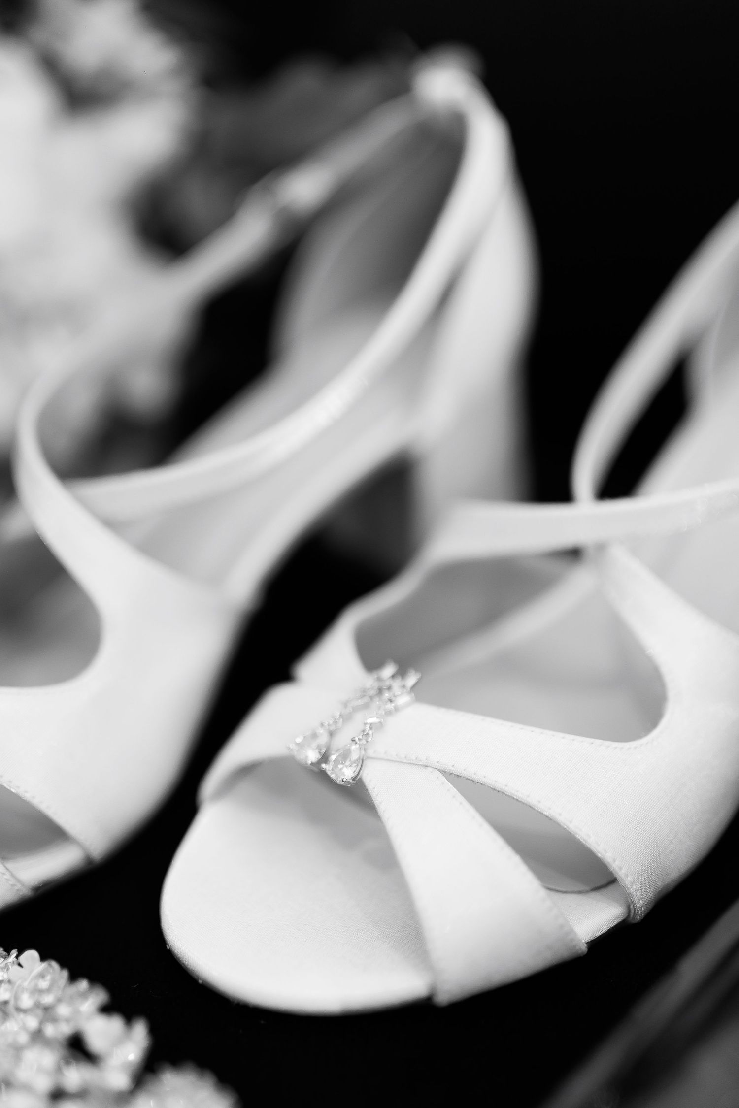 Black and white photo of elegant white bridal heels with crystal embellishment detail on a dark background.