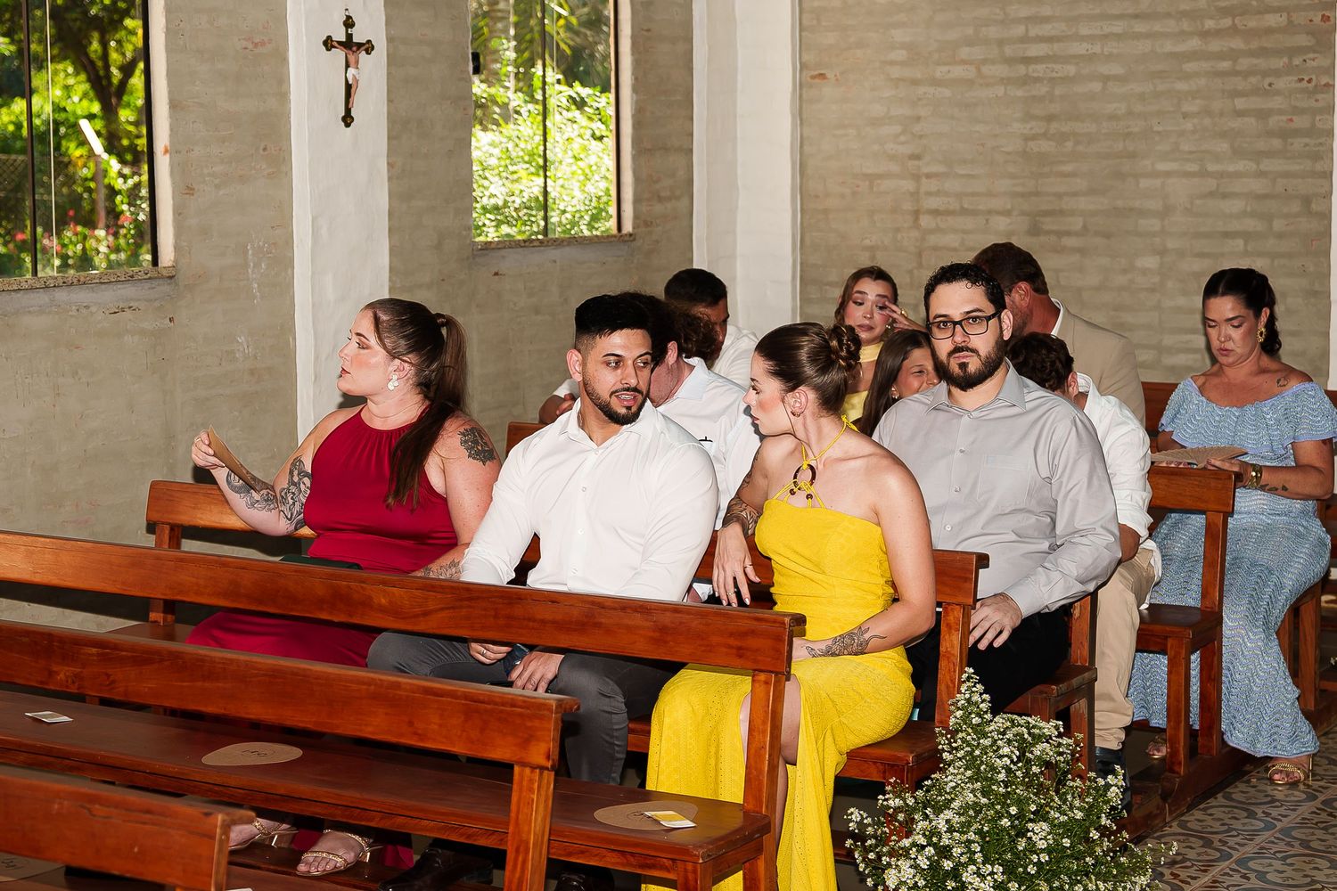 Wedding guests seated in wooden church pews, woman in yellow dress and man in red dress shirt among attendees.