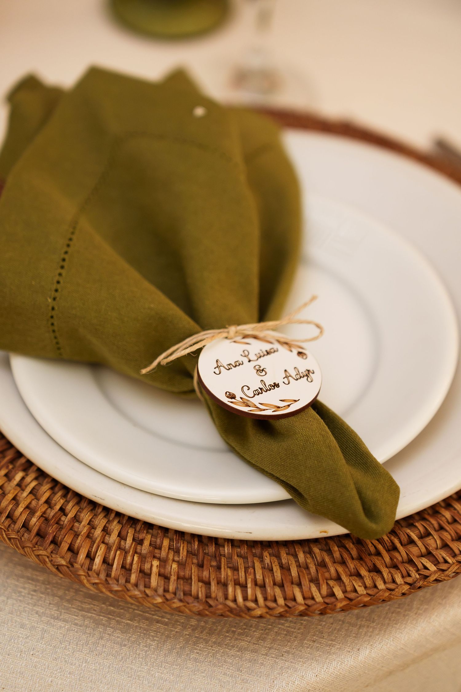 Elegant wedding place setting with olive green linen napkin, wooden name tag, white plates on woven charger.
