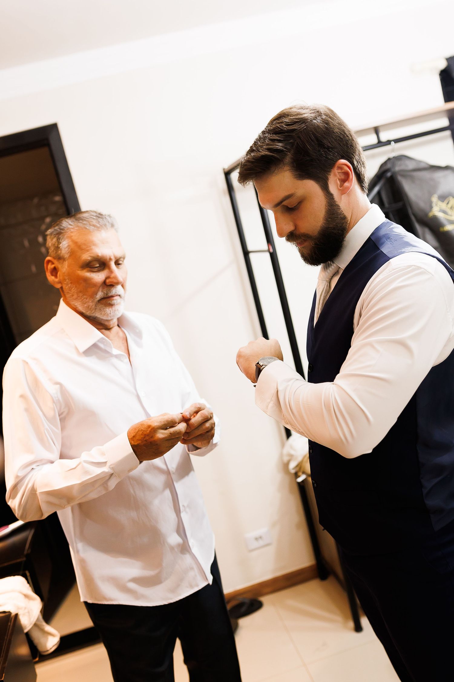 Two men in formal attire adjusting cufflinks while getting dressed for a special occasion indoors.