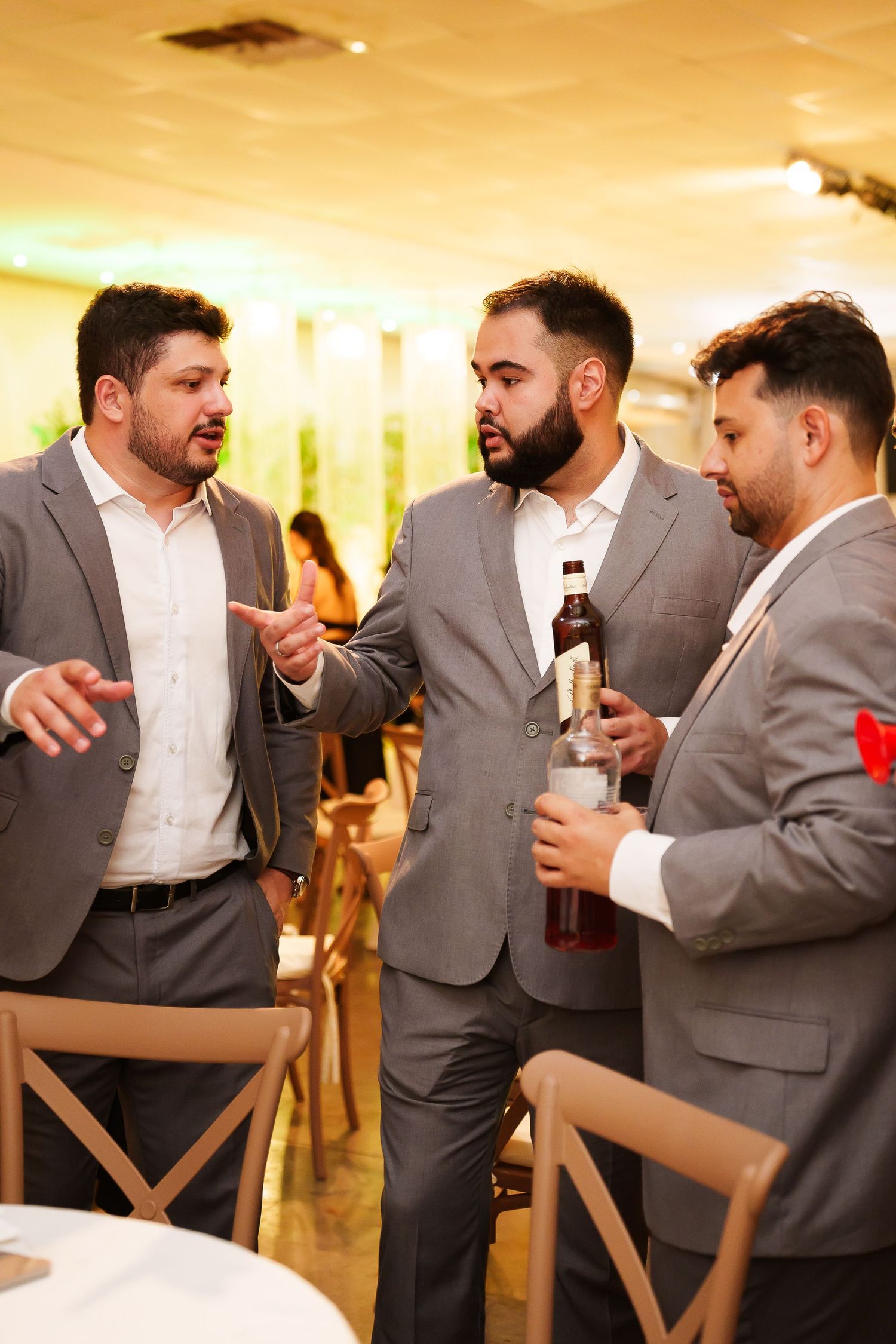 Three men in gray suits conversing at an indoor event, one holding a beer bottle.