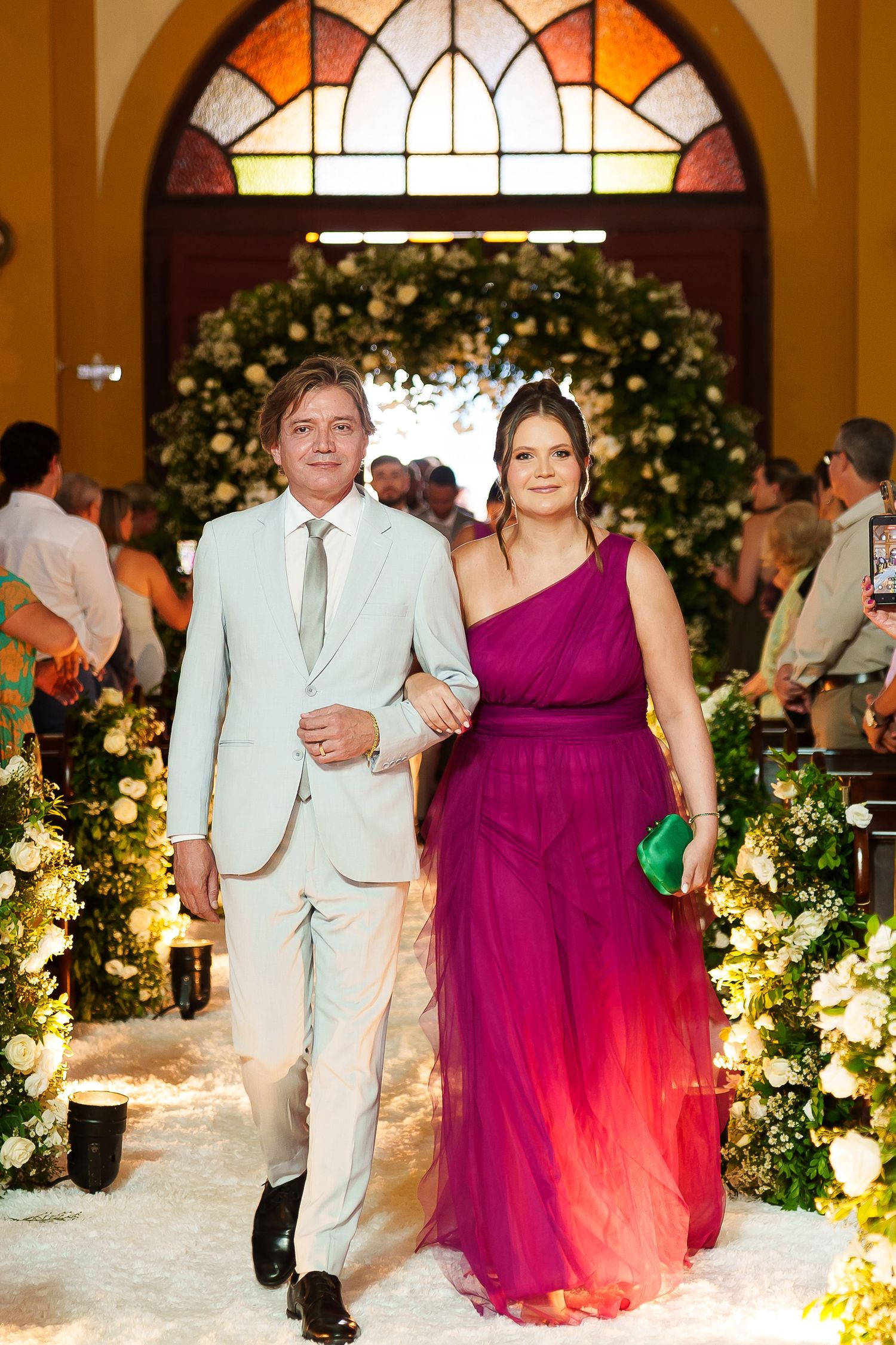 Elegant couple walking down a floral-decorated wedding aisle, man in light suit, woman in magenta ombre gown.