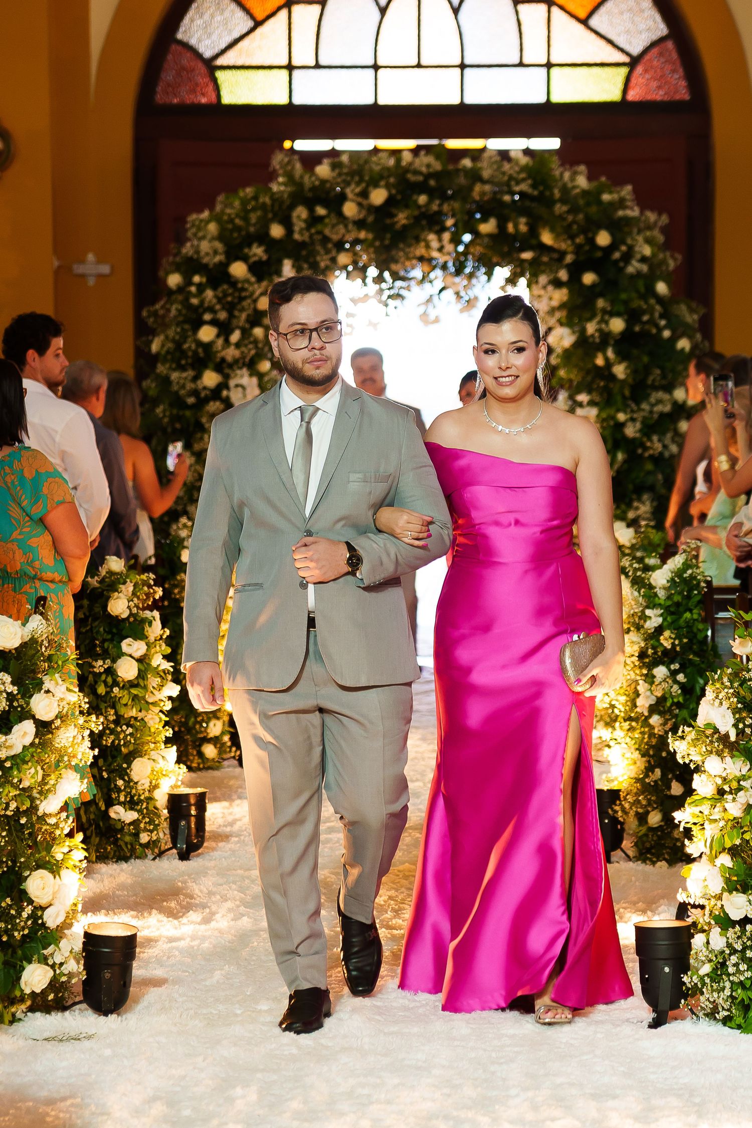 Couple walking down floral wedding aisle, man in gray suit, woman in pink satin gown.