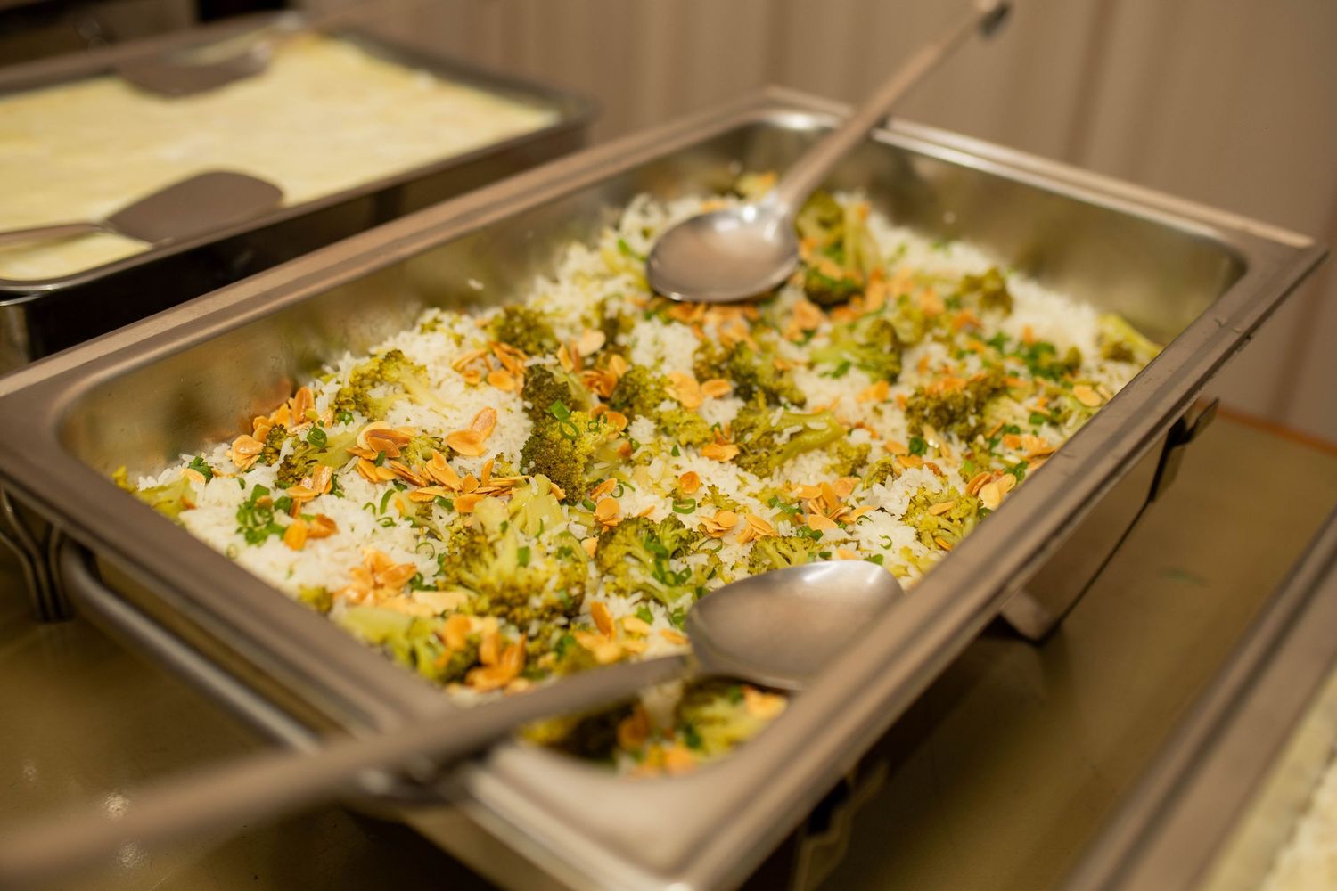 Buffet tray filled with vegetable fried rice featuring broccoli, carrots, and peas, served with large spoons.