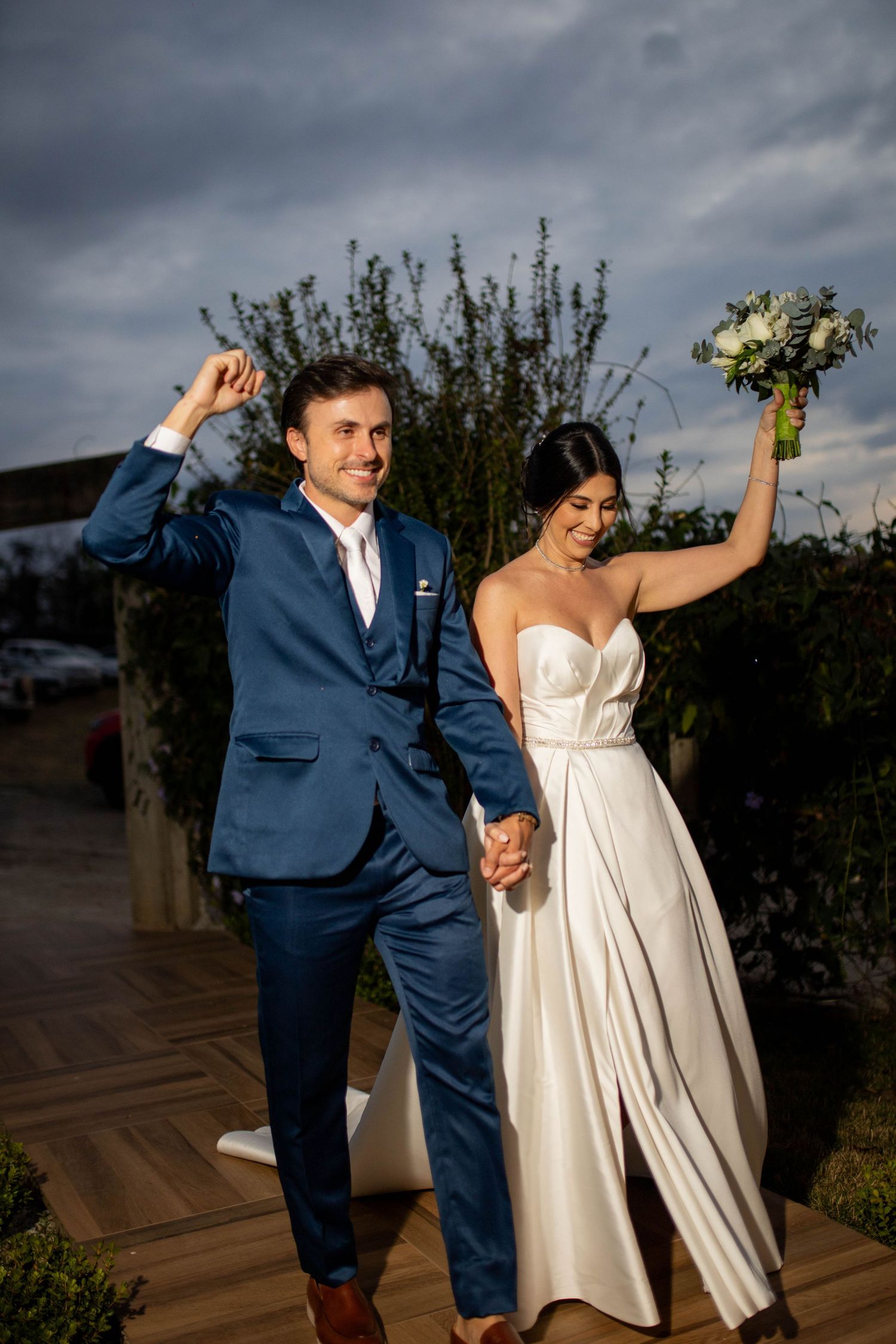 Joyful newlyweds celebrate outdoors, groom in blue suit, bride in white gown holding bouquet, dramatic cloudy sky backdrop.