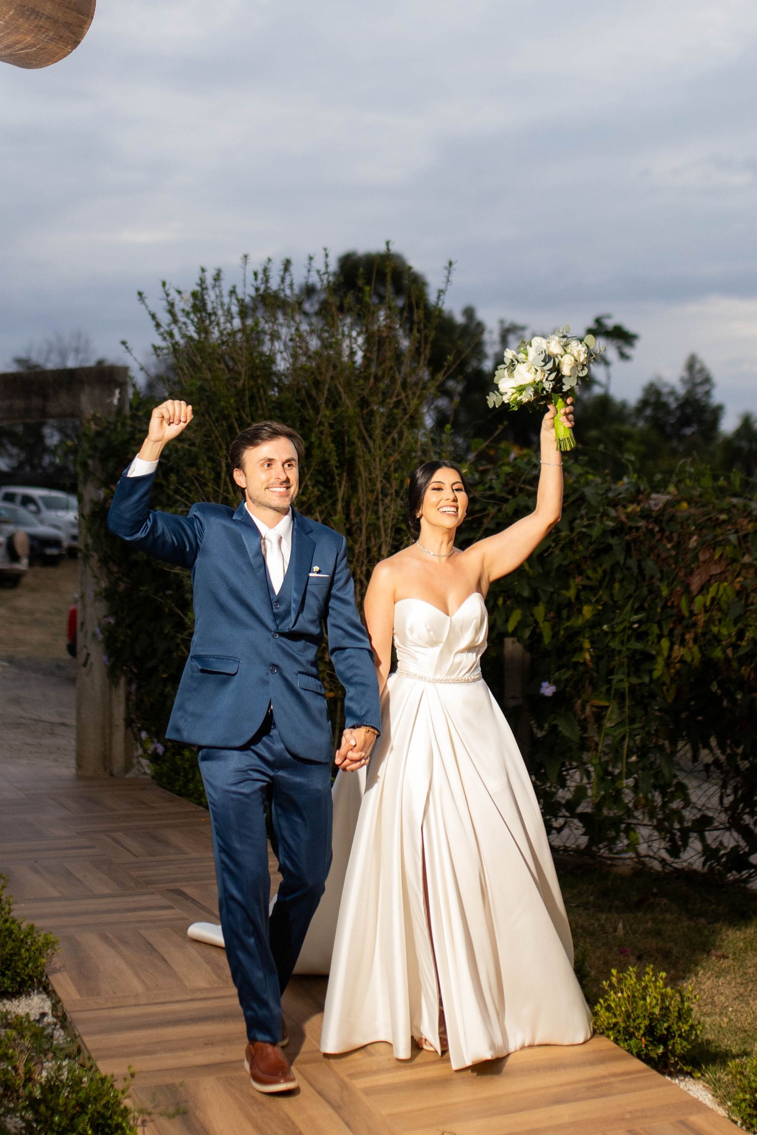 Joyful newlyweds celebrate outdoors, groom in navy suit raises fist, bride in white gown holds bouquet aloft.