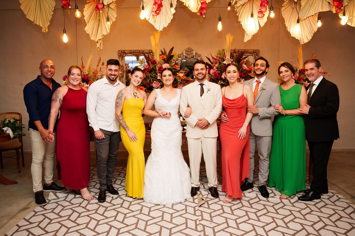 Wedding party posing together in colorful formal attire, bride in white gown, groom in beige suit, vibrant floral decor.